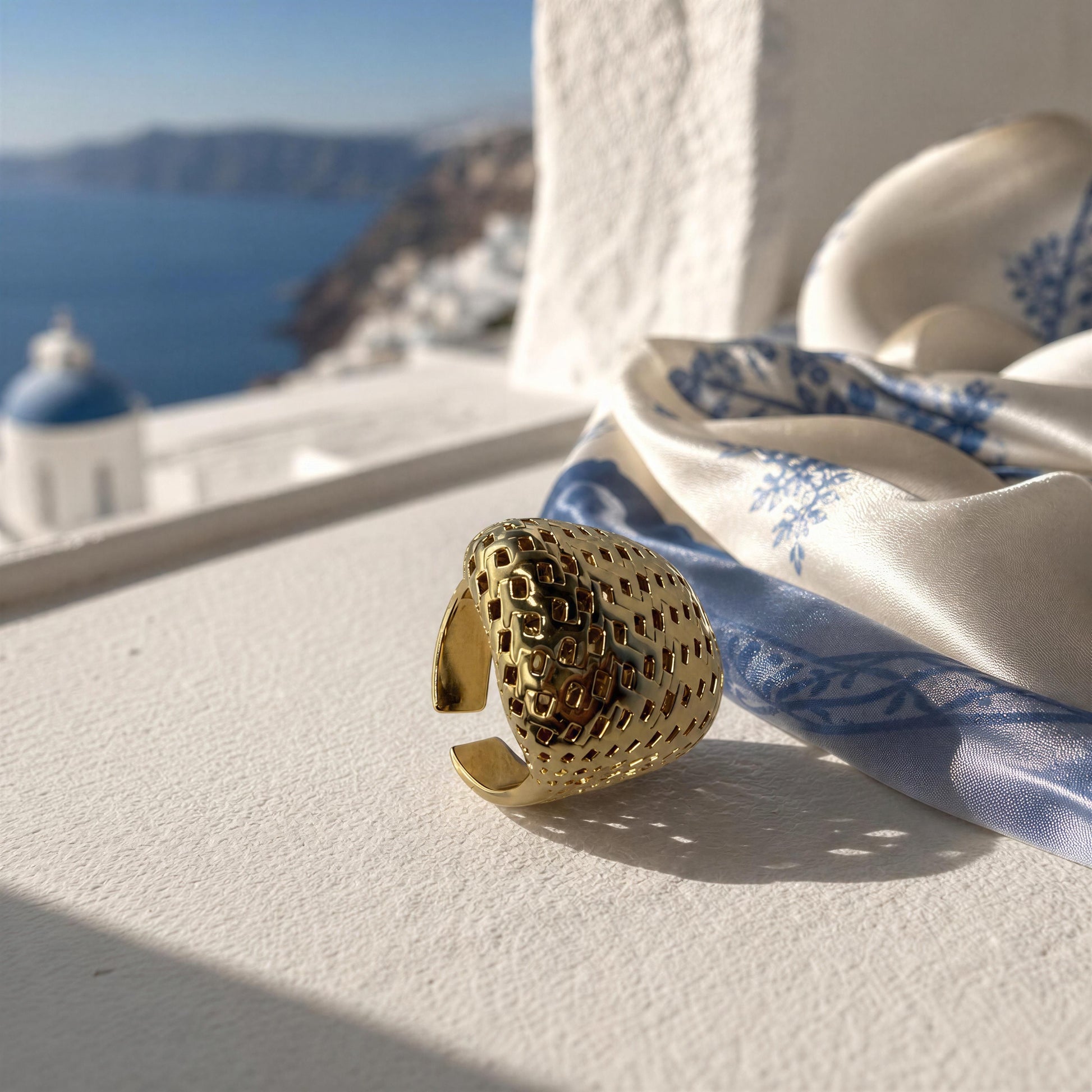 A textured gold ring with a honeycomb pattern, styled on a white surface next to a blue and white patterned silk scarf, with a blurred Santorini sea and white buildings in the background.