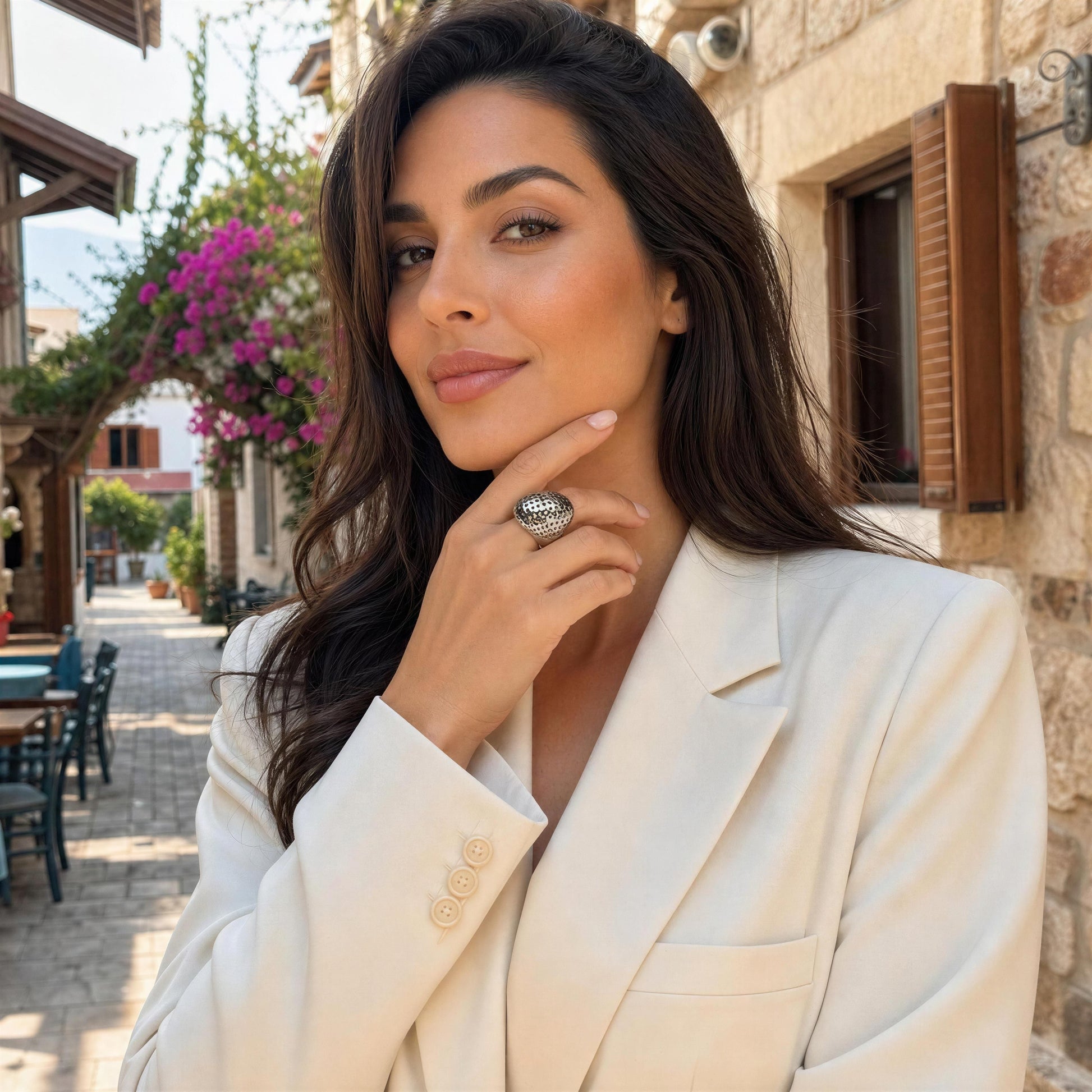 A woman wearing a cream-colored blazer and the Trama Ring from  Chica Chica Jewelry,  posing outdoors in a sunny stone street setting.