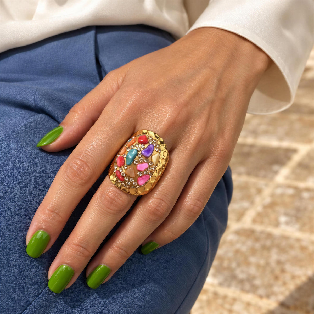 A close-up of a hand wearing the Chroma Ring - Chica Chica Jewelry, a large gold ring with colorful gemstones, styled with a white top and blue pants.