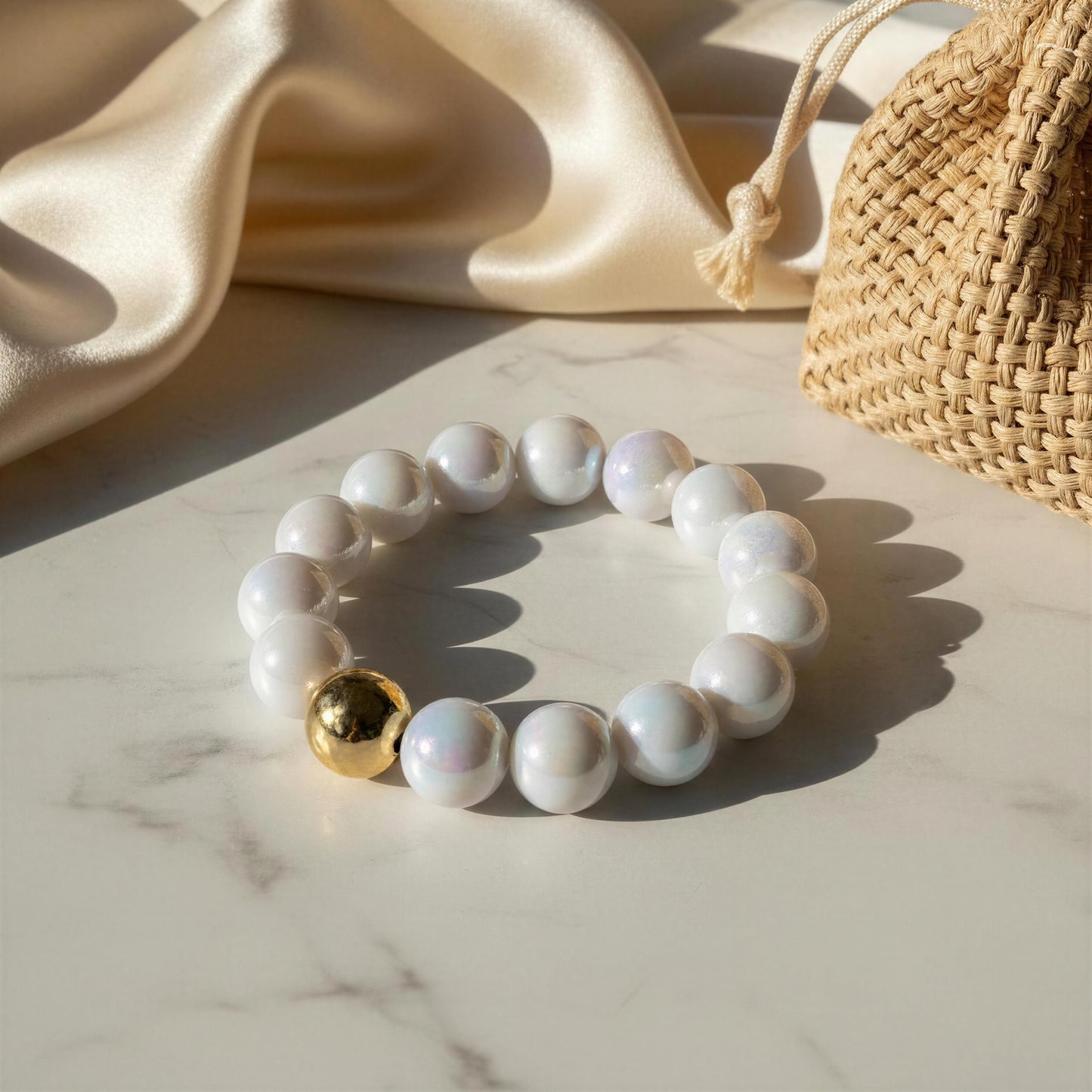 White beaded bracelet with a gold bead on a marble surface with a beige fabric and woven bag in the background.