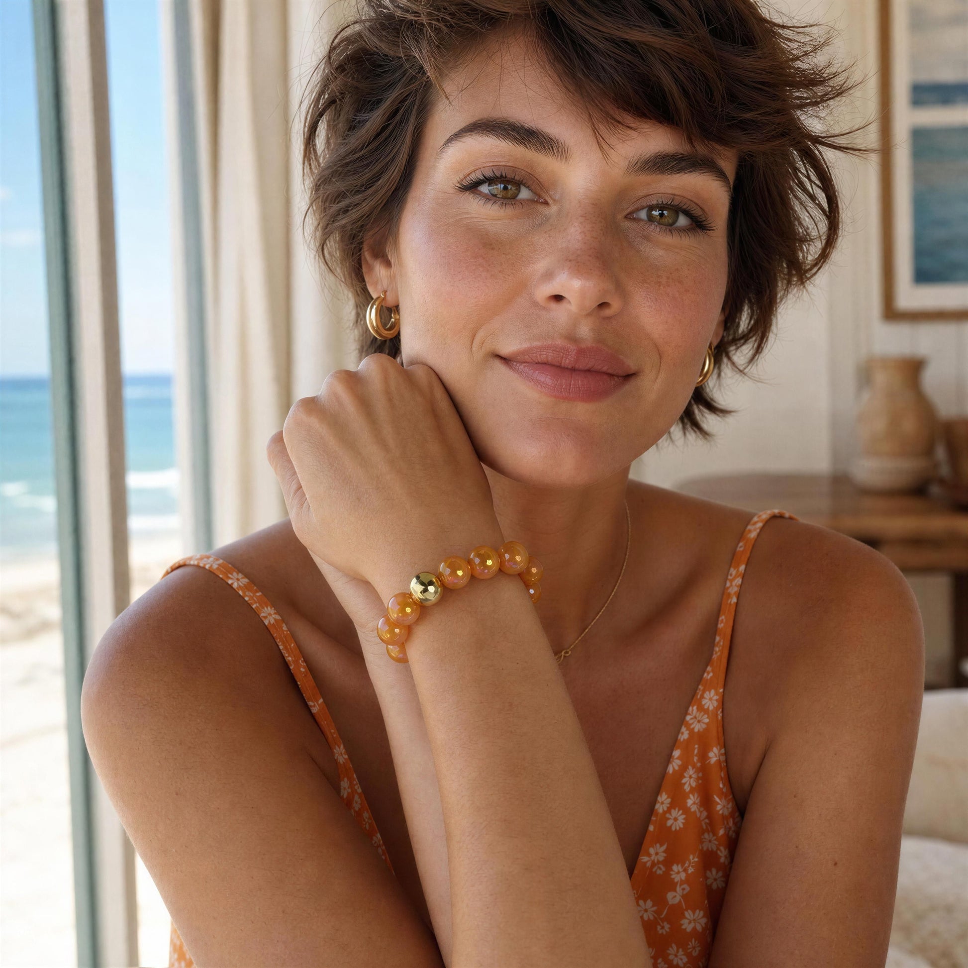 Woman with short hair wearing the Mandarin Glow Bracelet, sitting by a window with a view of the ocean.