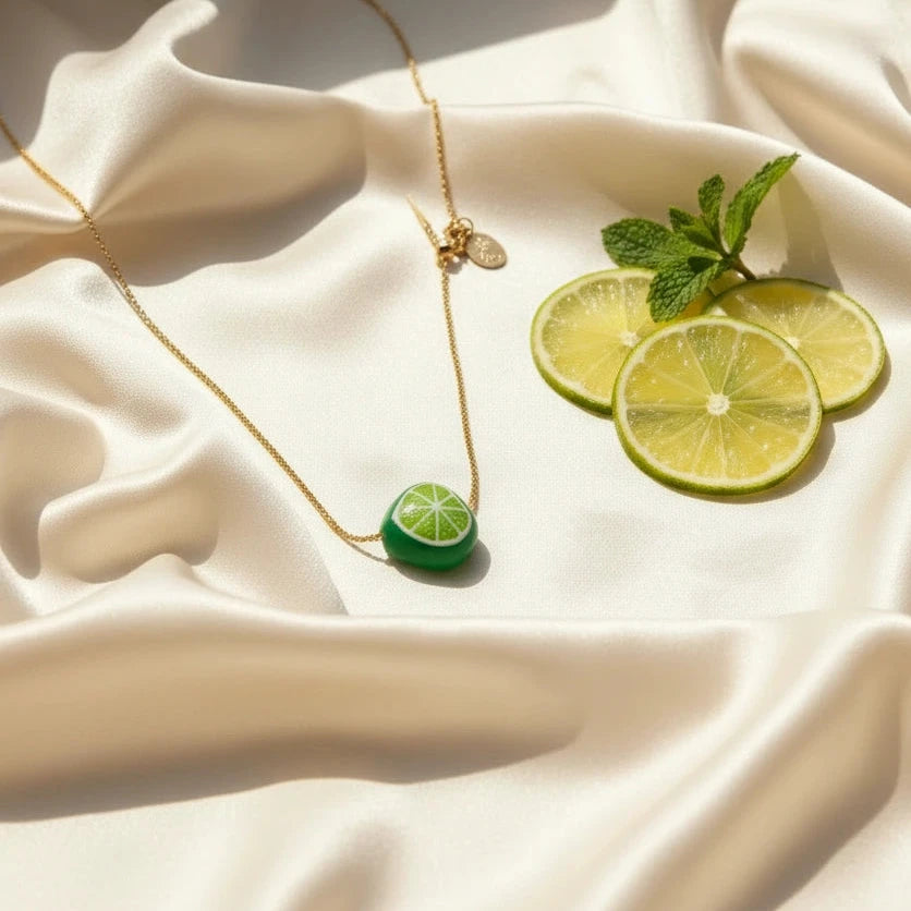 Tutti Frutti Lime Necklace with hand-painted green lime charm styled on soft fabric alongside fresh lime slices, showcasing vibrant color and gold-plated chain details.
