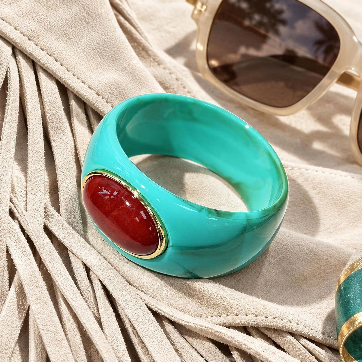 The Vittoria Bangle from  Chica Chica Jewelry; a  chunky turquoise bangle ring with a large oval burgundy wine cabochon and gold bezel, styled on a beige suede-like surface with fringe and sunglasses nearby.