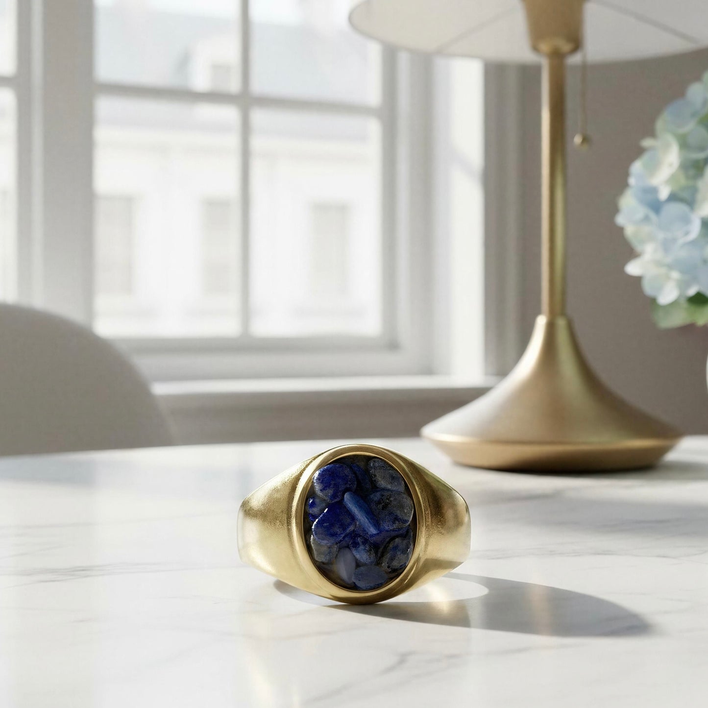 The Cosmic Blue Natural Stone Ring , displayed on a white marble tabletop in a softly lit room with a lamp and window in the background.
