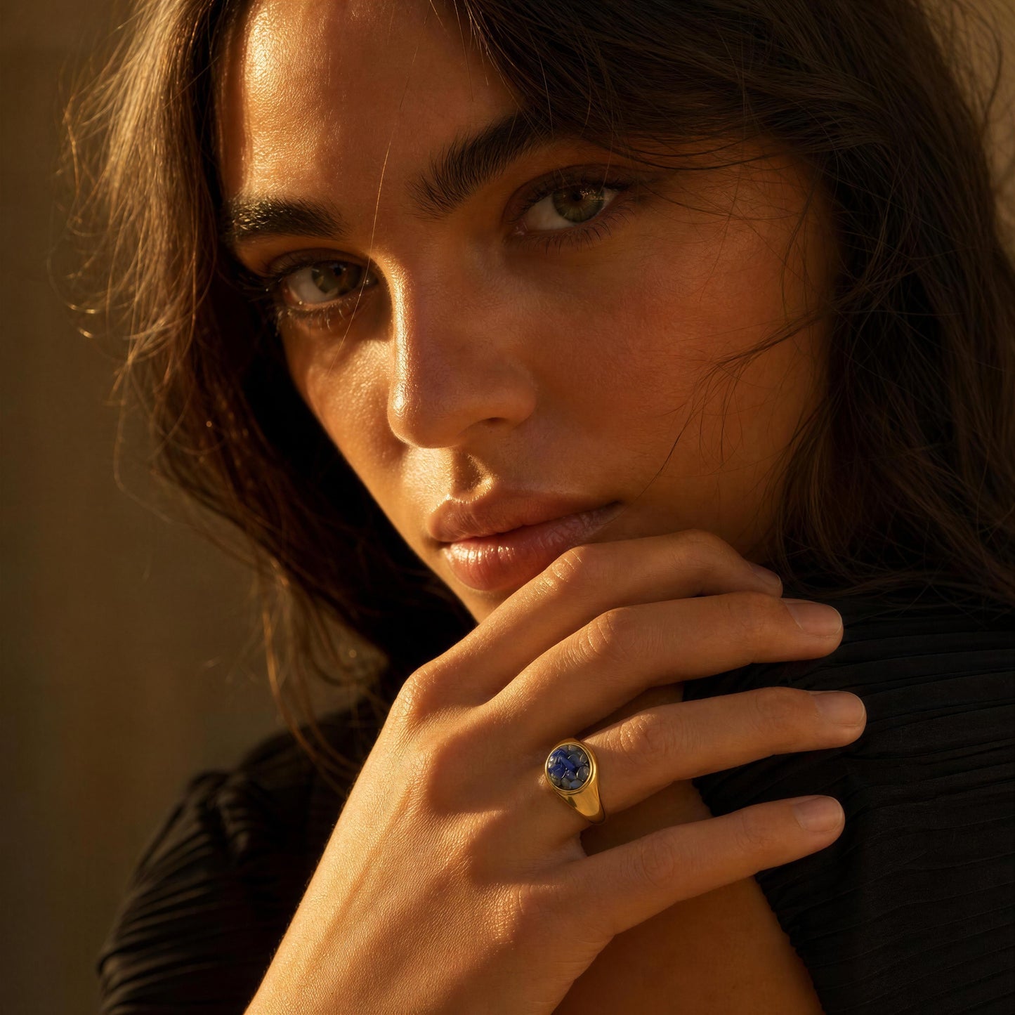 A close-up lifestyle shot of a woman wearing a gold ring with a deep blue oval gemstone, set against a warm, softly lit background.