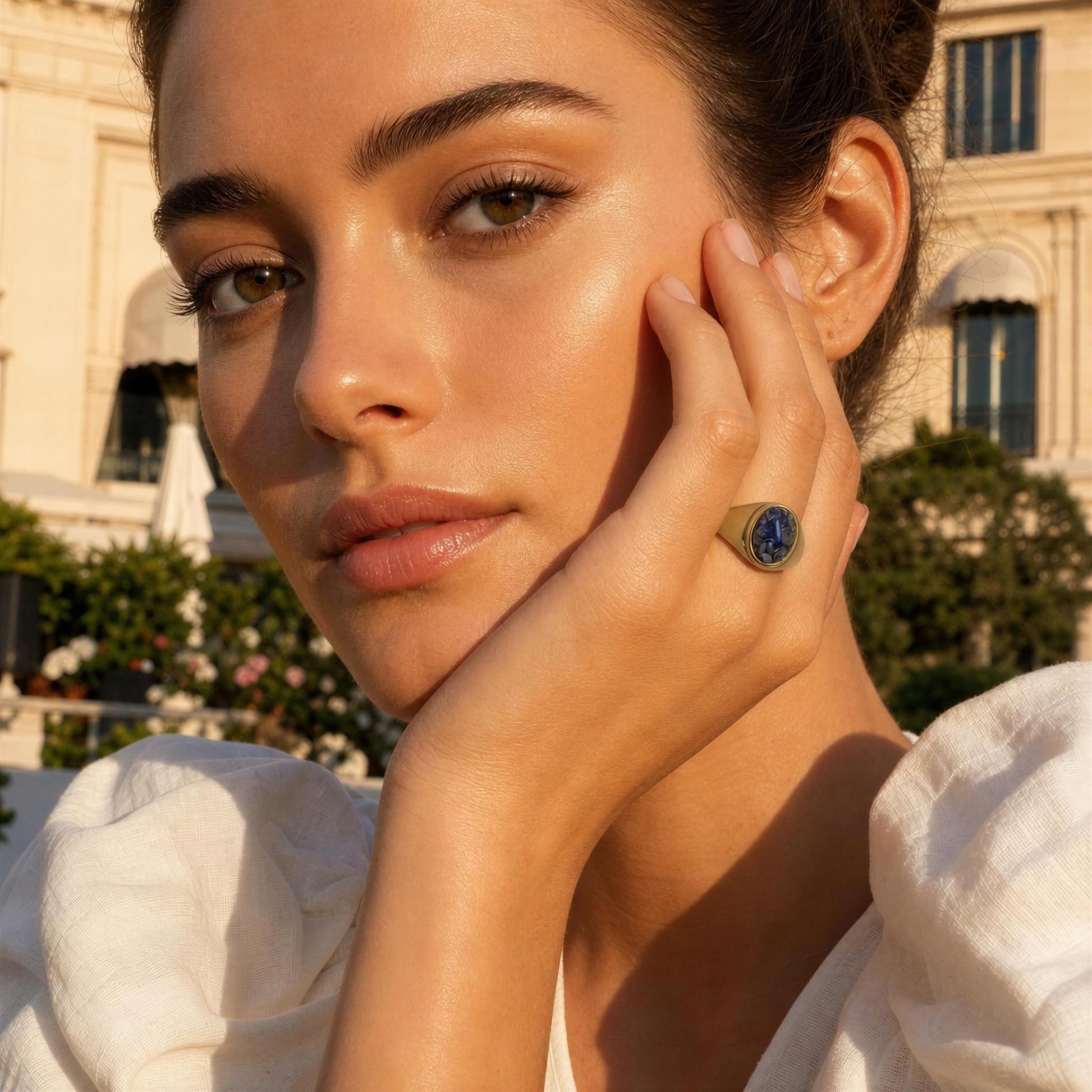 A close-up lifestyle shot of a woman wearing the Cosmic Blue Natural Stone Ring,  gold ring with a blue stone, posed outdoors against a warm, sunlit background with a classical building.