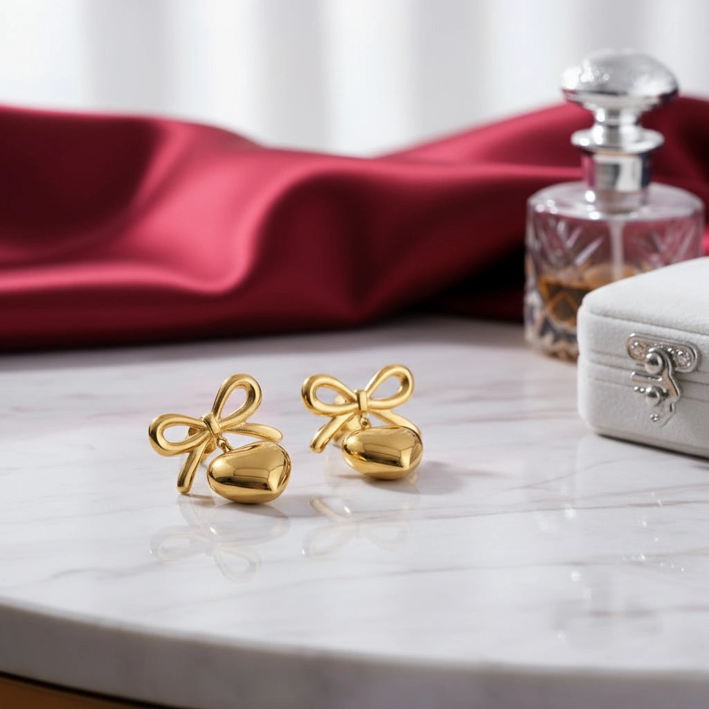 Gold bow-shaped earrings on a marble surface with a red fabric and perfume bottle in the background.