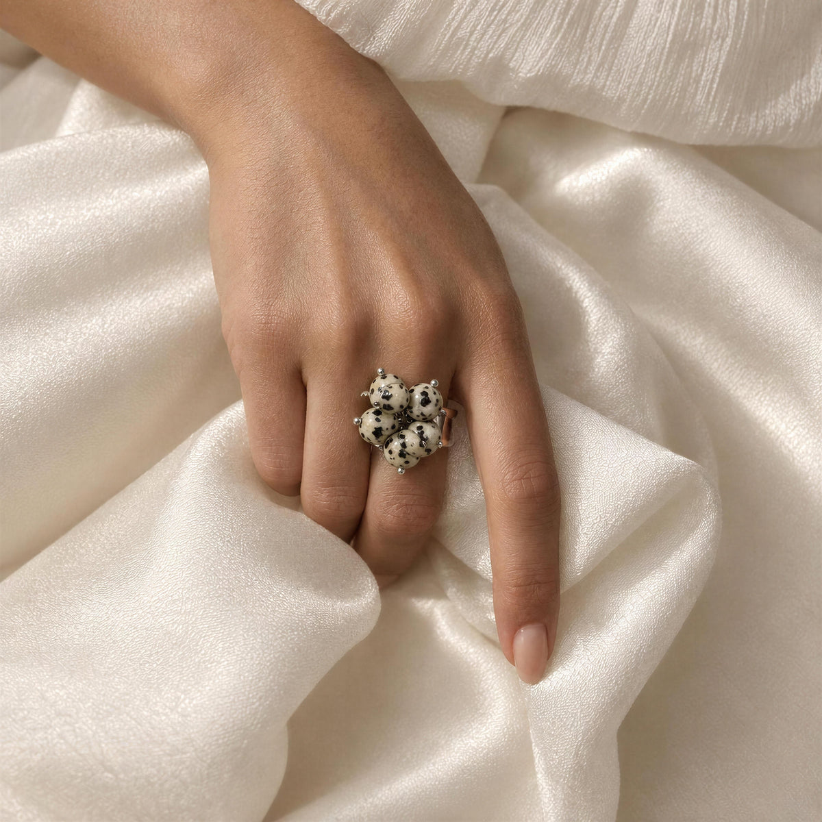 A close-up of a hand wearing the Dalma Ring from Chica Chica Jewelry-  a unique ring with four black-and-white spotted dalmatian-style faces, resting on draped white satin fabric.