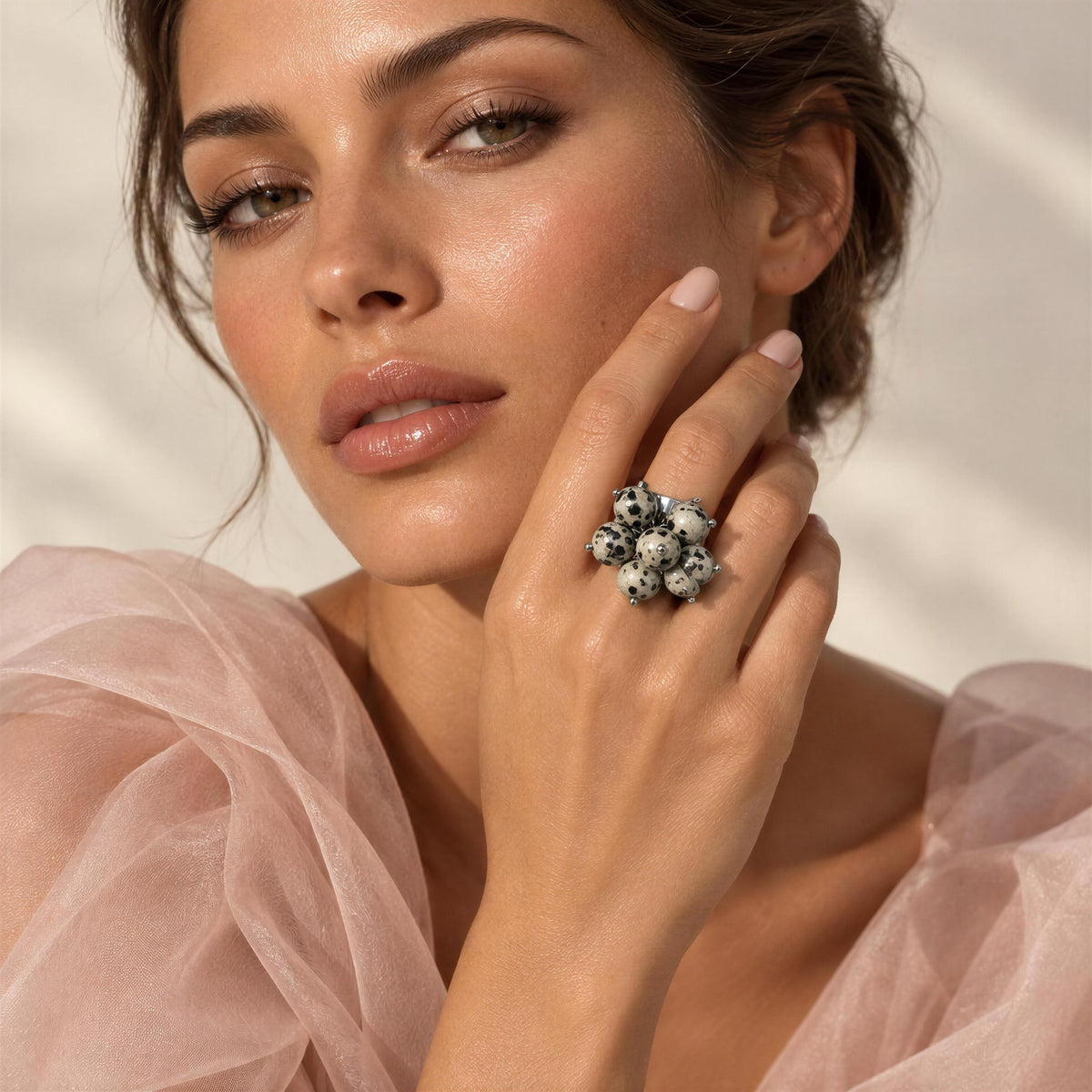 A close-up lifestyle shot of a model wearing the Dalma Ring from Chica Chica Jewelry,  a large, chunky ring made of black and white marbled stone clusters on her finger, paired with a light pink tulle top against a soft beige background.