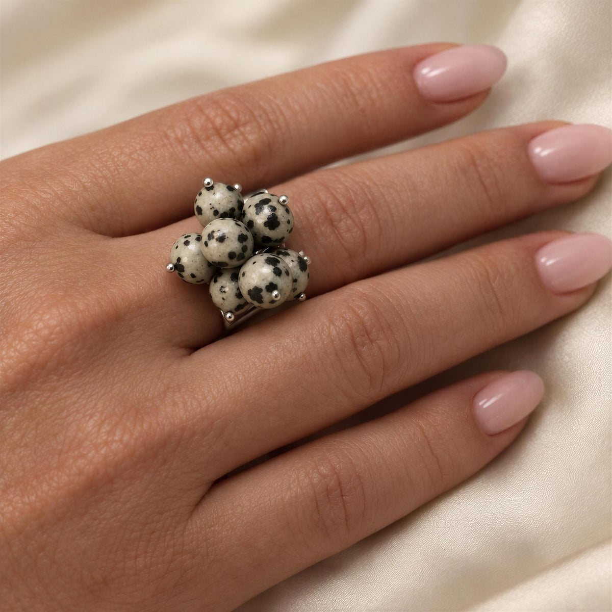 A close-up of a hand wearing a cluster ring made of white and black spotted beads on a silver band, set against a soft cream-colored fabric background.