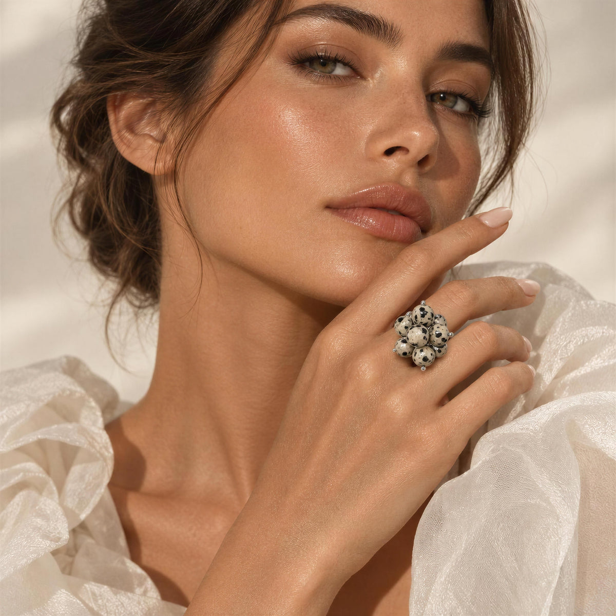 A close-up lifestyle shot of a woman wearing a large, ornate silver ring with a floral cluster of clear stones. She is wearing a white ruffled blouse against a softly lit, warm-toned background.