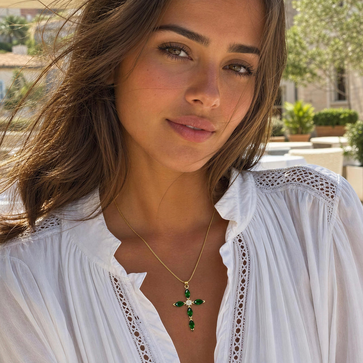 A close-up lifestyle shot of a woman wearing a gold cross pendant necklace with green emerald-cut stones and a small white crystal accent, paired with a white blouse.