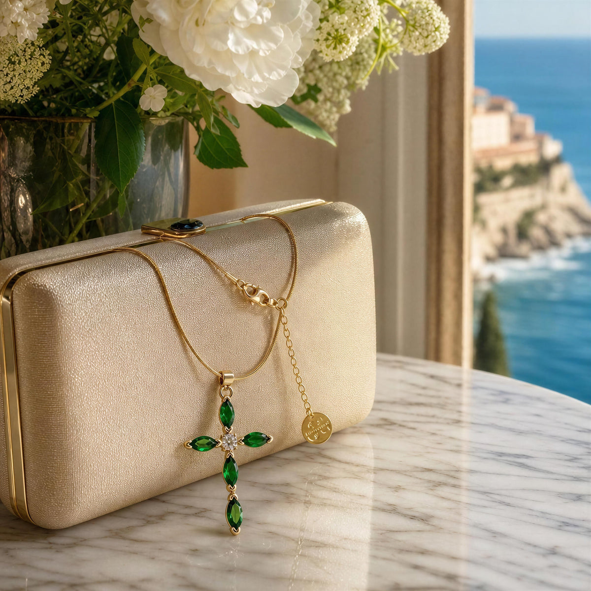 A gold cross pendant with green marquise-cut stones and a small round gold coin charm, draped over a champagne clutch on a marble table with a coastal view in the background.