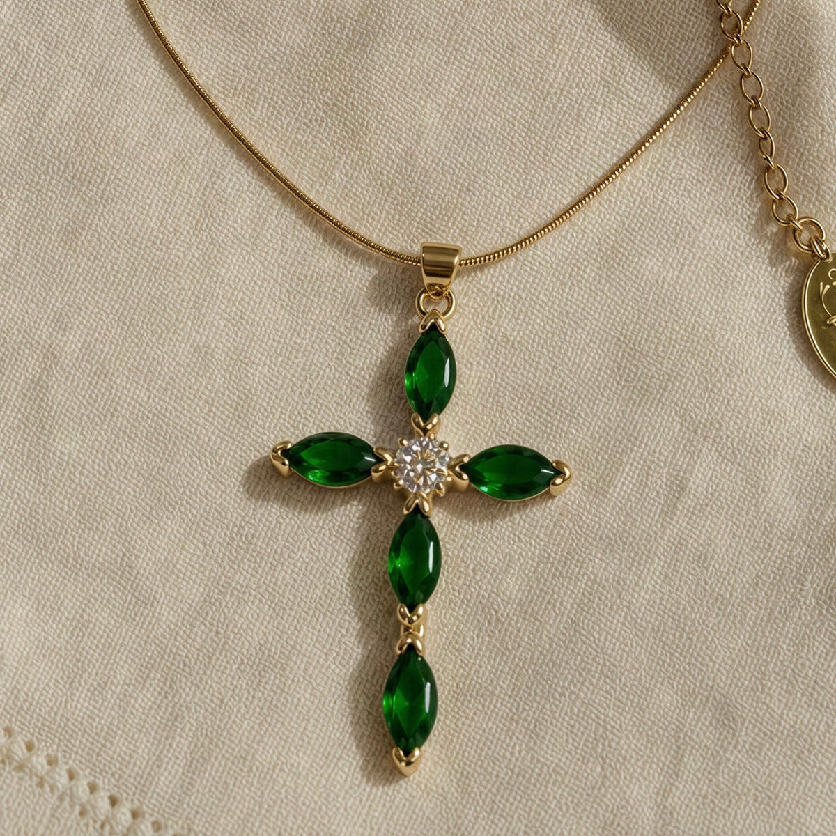 The Defne Cross Necklace from Chica Chica Jewelry; a gold  cross pendant necklace featuring four green marquise-cut stones and a small clear crystal center, displayed on a textured beige fabric background.