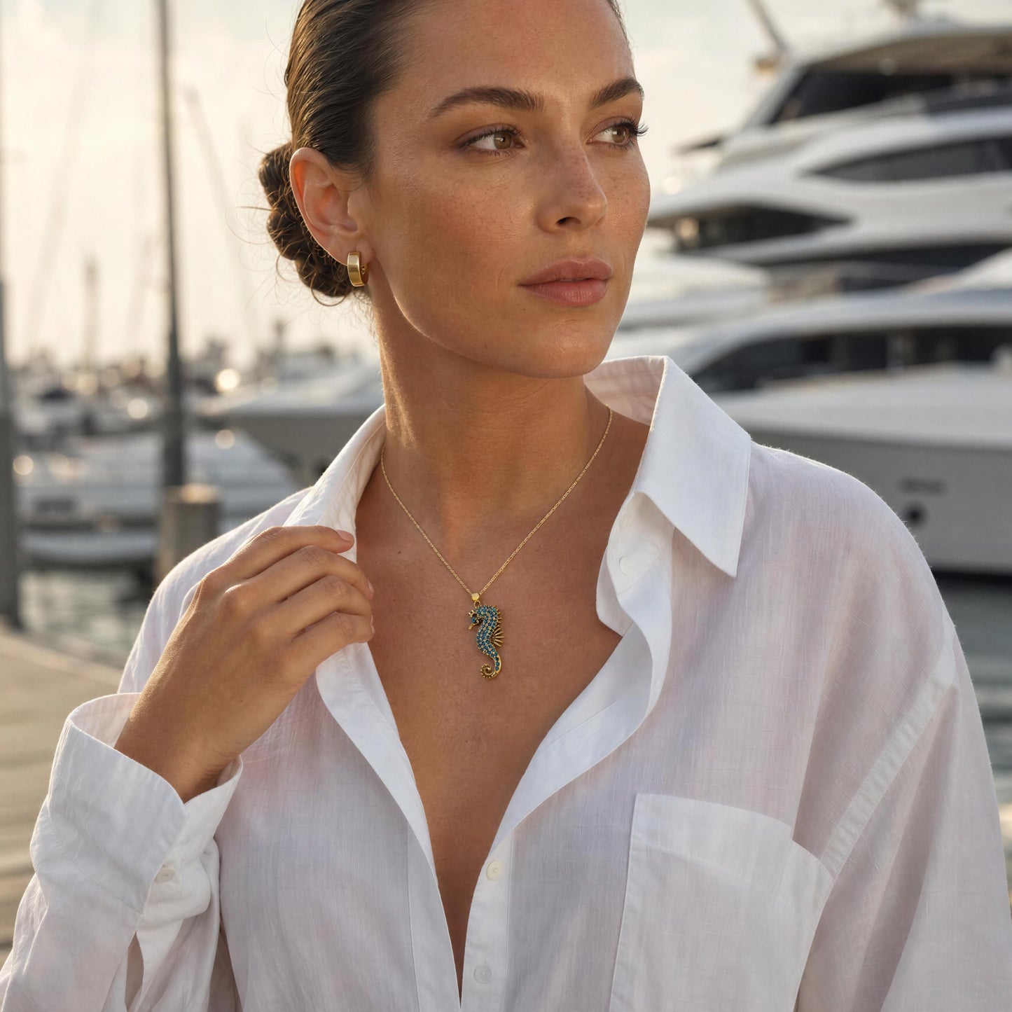 A woman wearing a delicate gold necklace with a seahorse pendant, standing in a marina with luxury yachts in the background. She is wearing a white button-down shirt.
