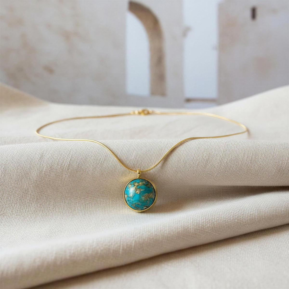 Close-up of Derya Necklace featuring a round turquoise blue pendant with gold veining and gold-tone setting on vainilla color fabric. 