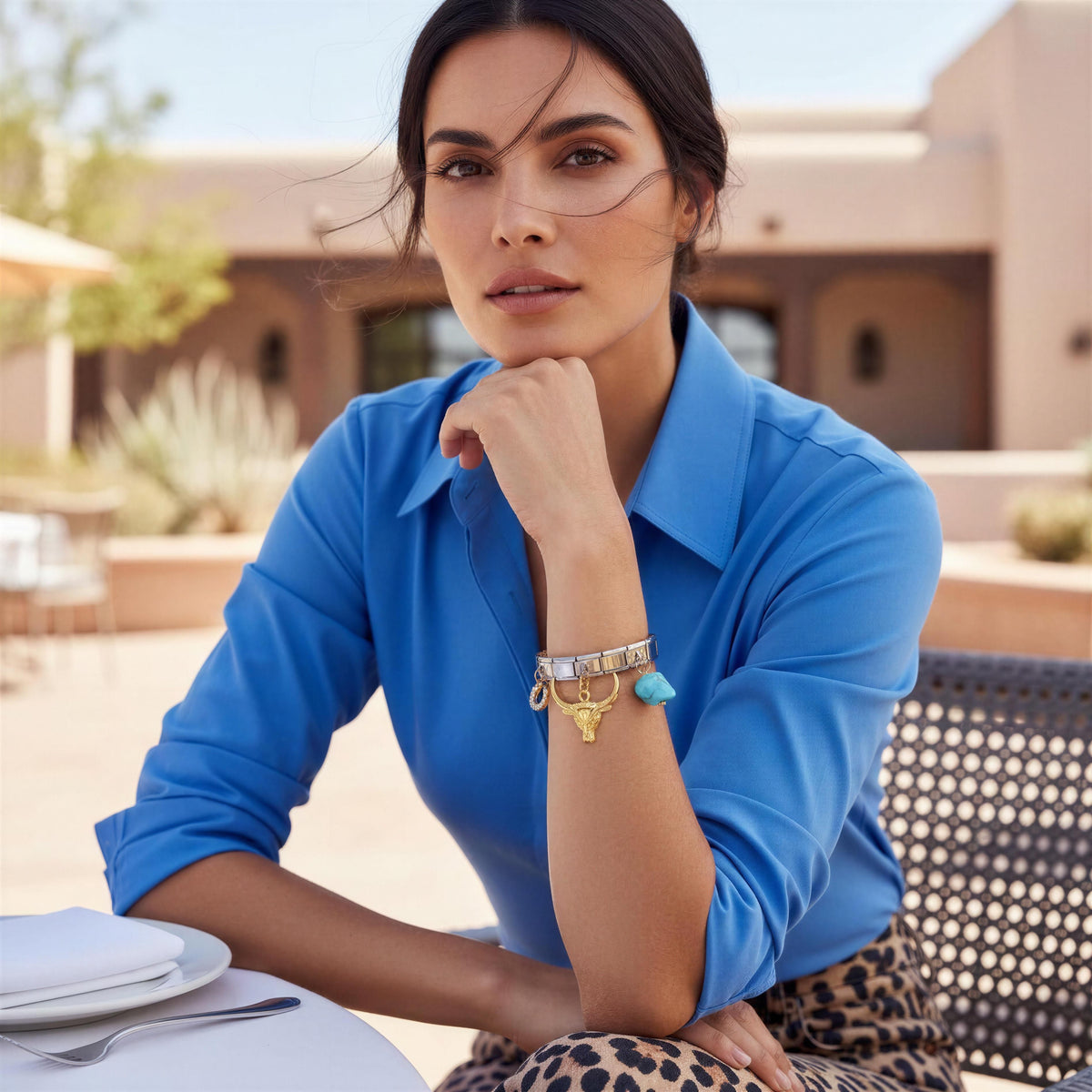 A woman wearing a blue button-up shirt and leopard-print bottoms sits at an outdoor table, wearing a silver charm bracelet with gold and turquoise details on her wrist.