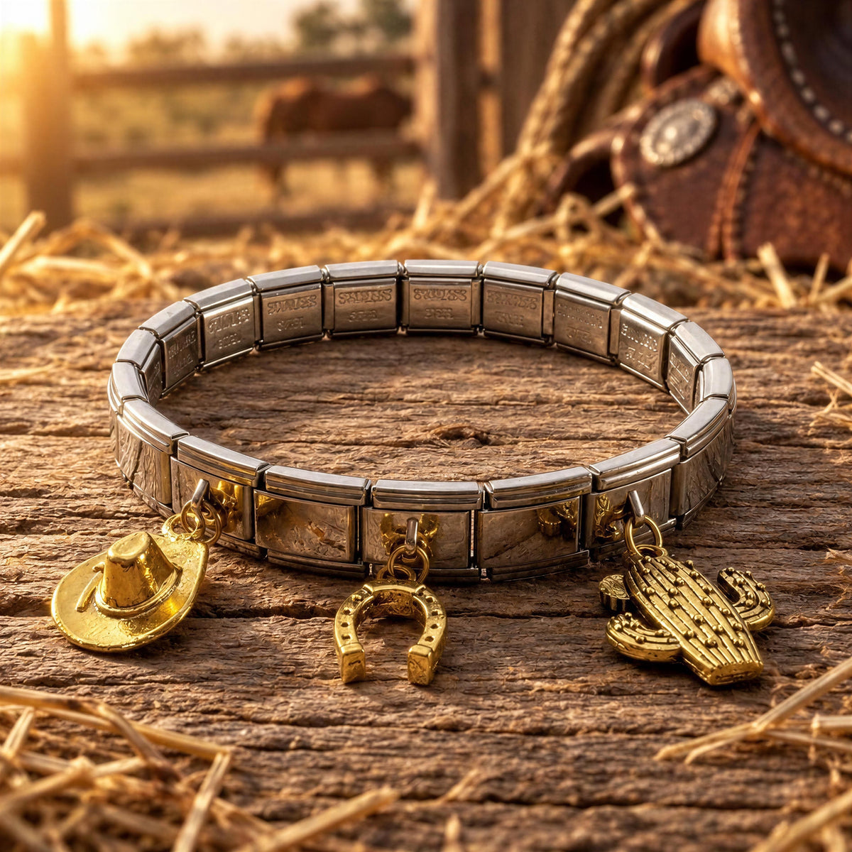 A silver-toned stretch bracelet with rectangular links and three dangling gold-tone charms shaped like a cowboy hat, horseshoe, and cactus, displayed on a rustic wooden surface with straw and a blurred outdoor ranch background.