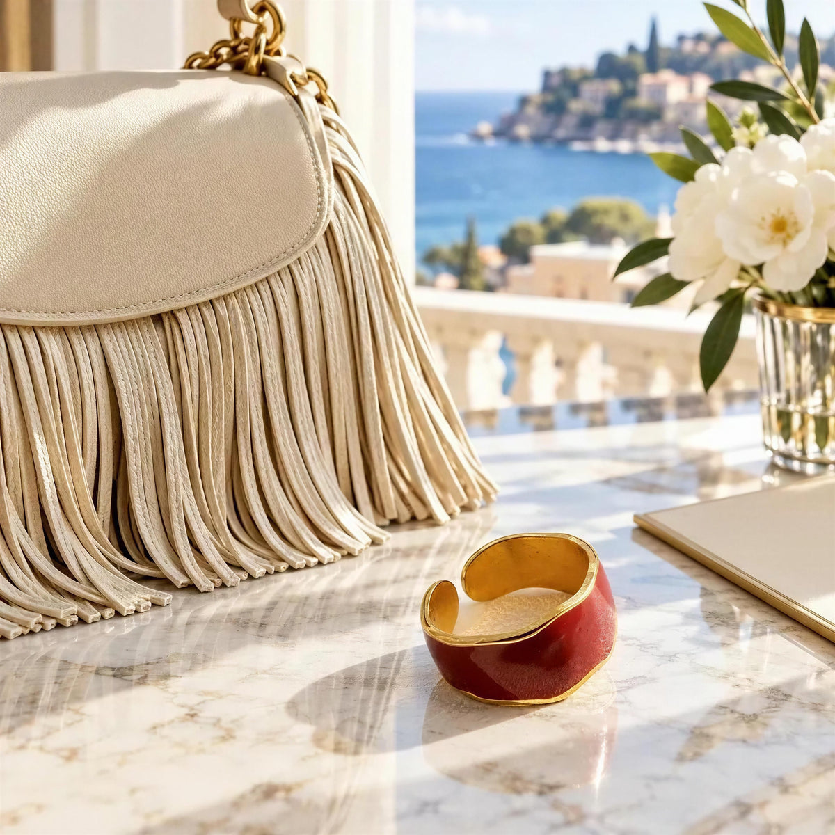 The Duna Ring – Carmine from Chica Chica Jewelry on marble desk next to a cream color fringes handbag with a view of the Mediterranean. 