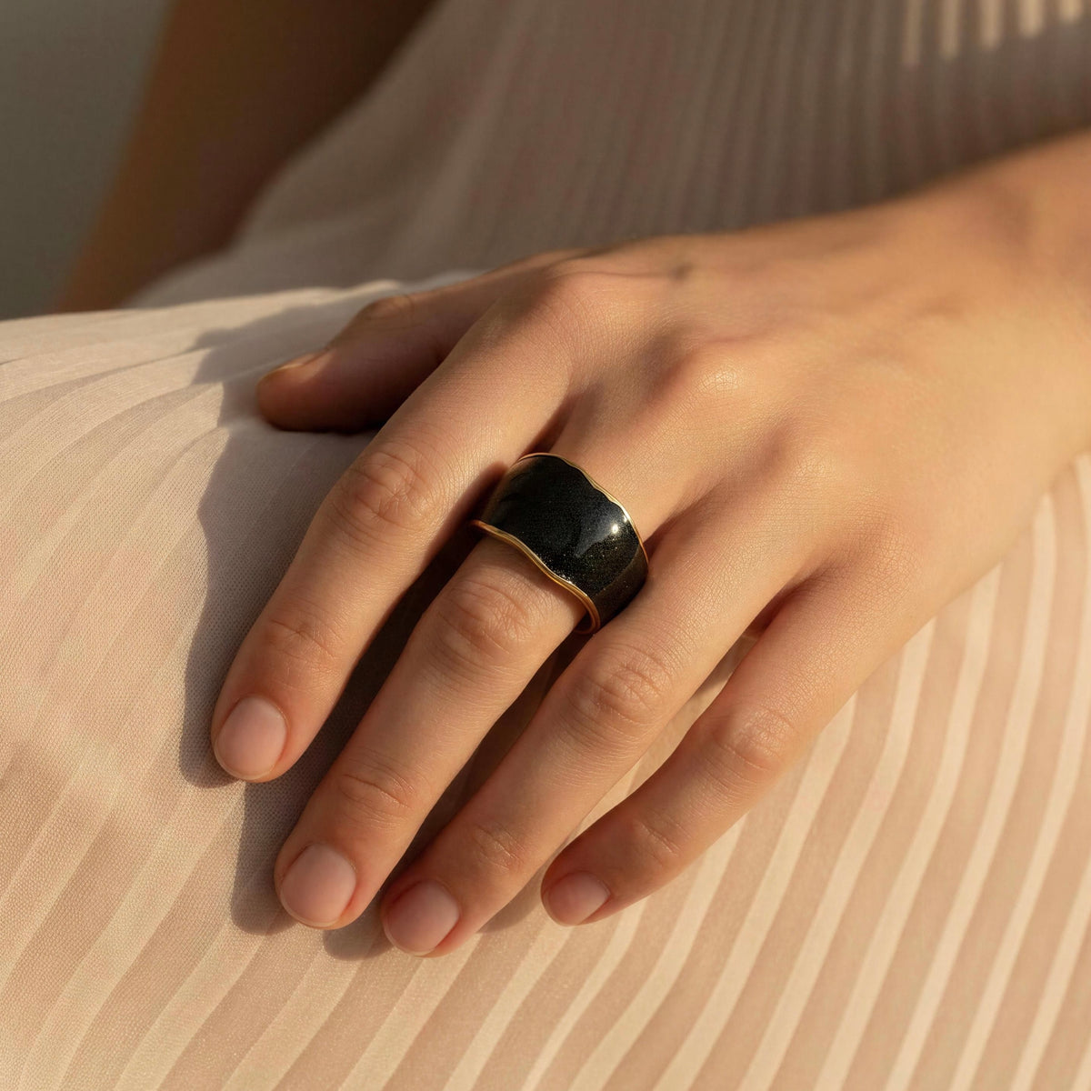 A close-up of a hand wearing the Duna Ring – Ónice from Chica Chica Jewelry, a wide black ring with a gold-toned band, resting on a beige ribbed fabric surface.