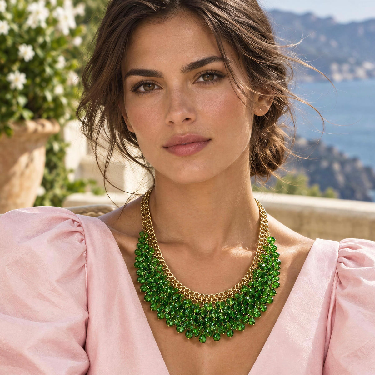 A woman wearing the Eleonora Necklace
from Chica Chica Jewelry, a chunky statement necklace made of layered green faceted beads and gold chains, paired with a light pink blouse, set against a scenic outdoor background with a stone wall and blue sea.
