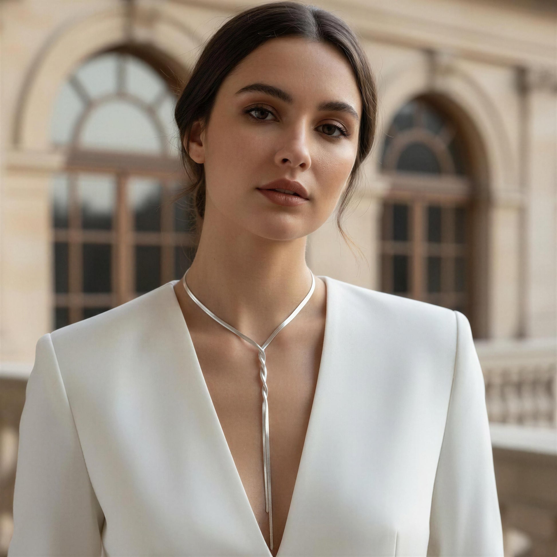 Woman wearing a white outfit with a deep neckline and the Eterna Lariat necklace, standing in front of an architectural background.
