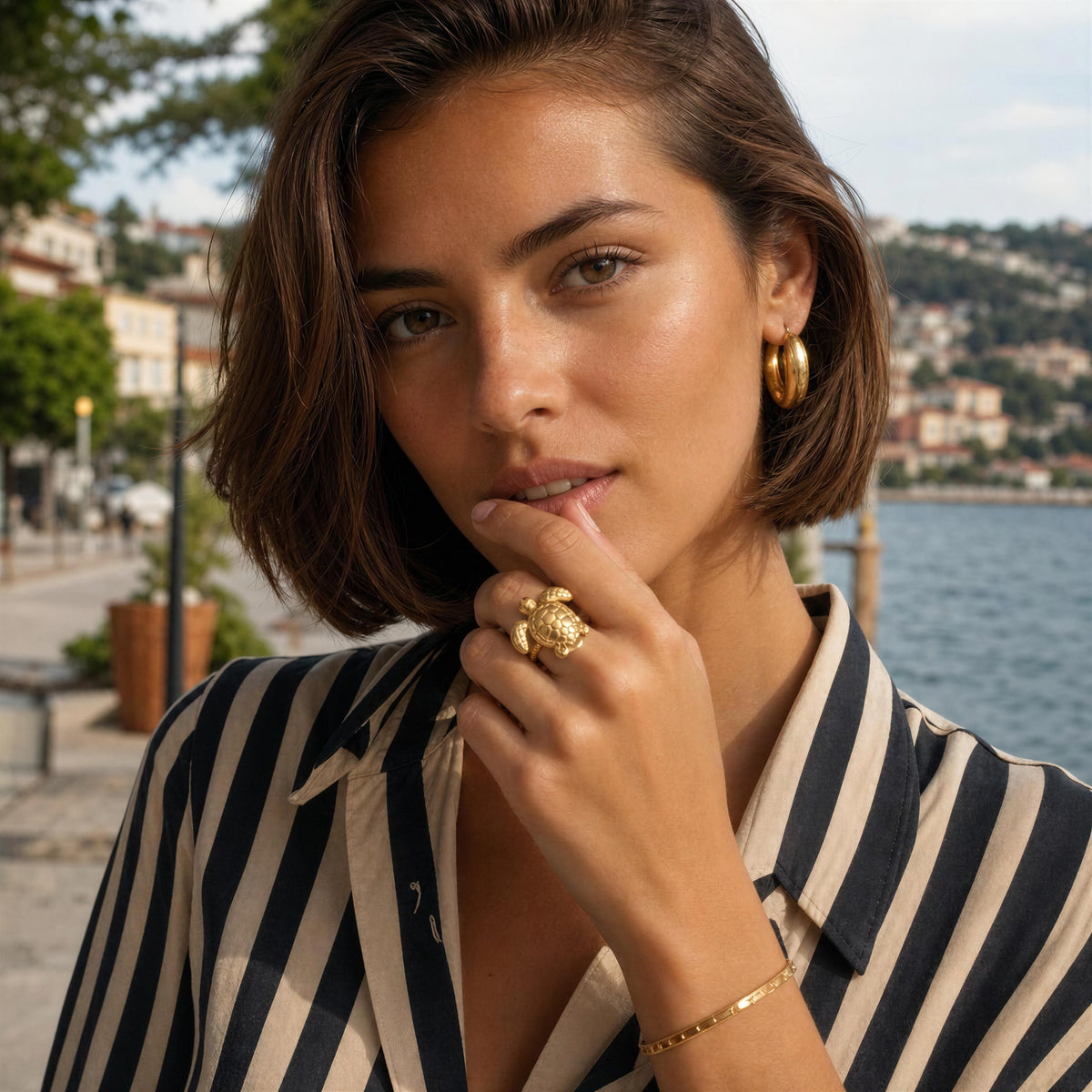 A woman with short brown hair poses outdoors near the water, wearing a  turtle gold ring on one finger and a gold hoop earring. She is wearing a beige and navy vertically striped button-up shirt.