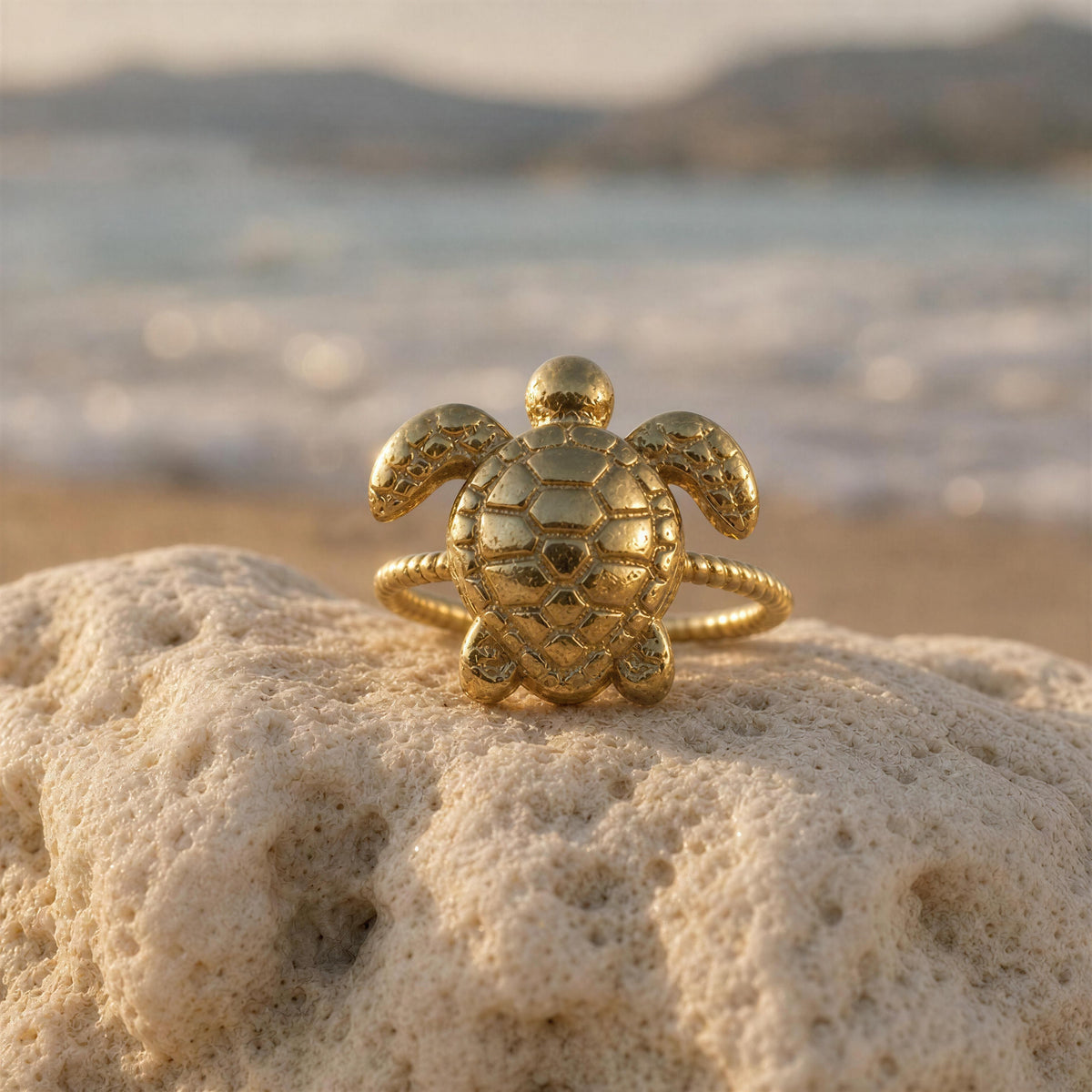 The Eternal Tortoise Ring from Chica Chica Jewelry, a gold turtle-shaped ring with a textured, scale-like shell sits on a light beige rock with a blurred beach and ocean background.