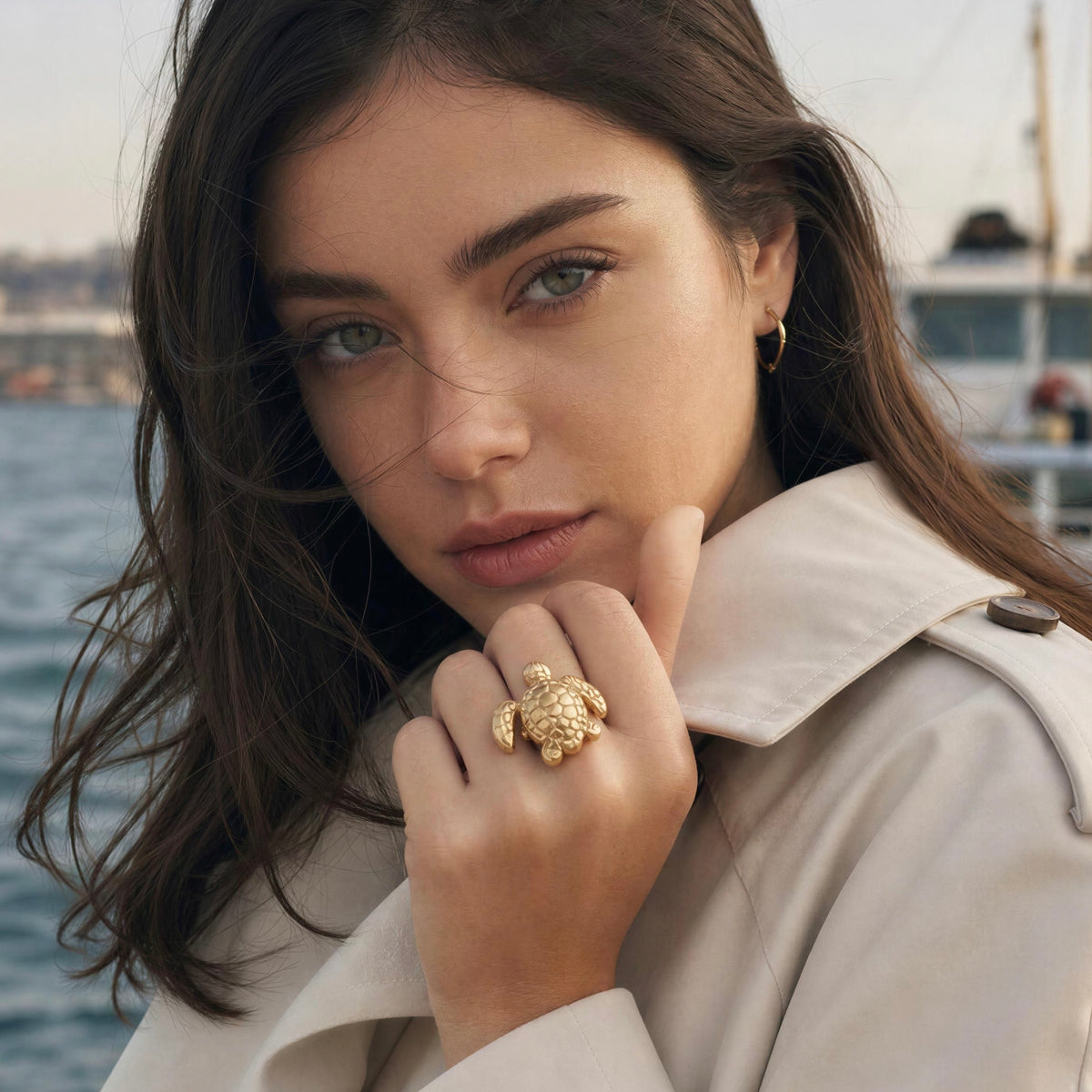A close-up lifestyle shot of a woman with long dark brown hair wearing a beige trench coat, featuring a prominent gold turtle ring on her finger, with a blurred harbor background.