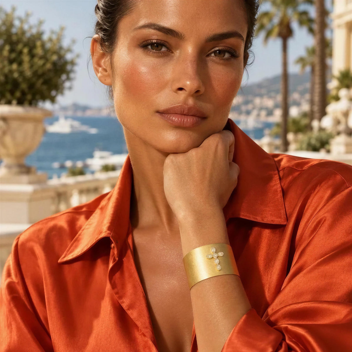 A close-up lifestyle shot of a woman wearing a wide, polished gold cuff bracelet with small white pearl-like accents. She is wearing a vibrant orange satin shirt, with a sunny coastal resort background featuring blue water, white yachts, and palm trees.