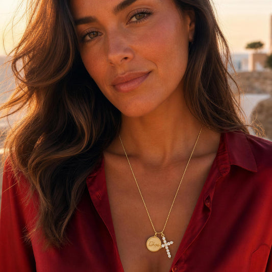 A woman wearing the Fede Necklace from Chica Chica Jewelry , a delicate gold necklace with a round Dios medallion and a small cross pendant, styled with a red satin shirt against a warm, blurred outdoor background.
