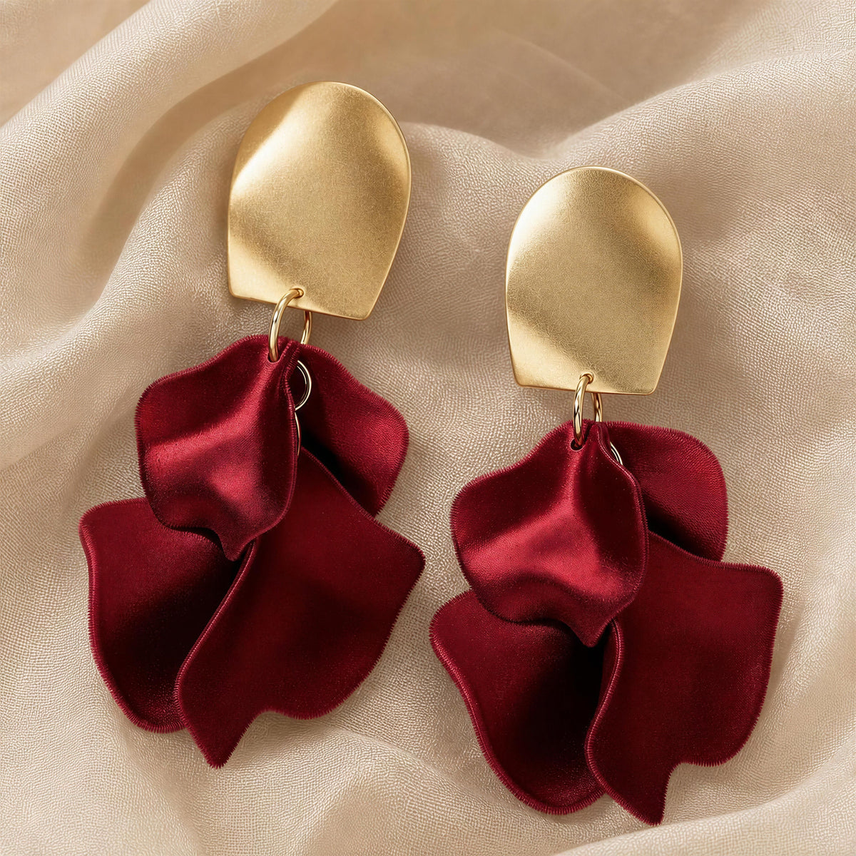 The Fiore Roja earrings, a pair of statement dangle earrings with textured gold-tone metal top pieces and layered deep burgundy satin petal charms, displayed on a draped beige fabric background.