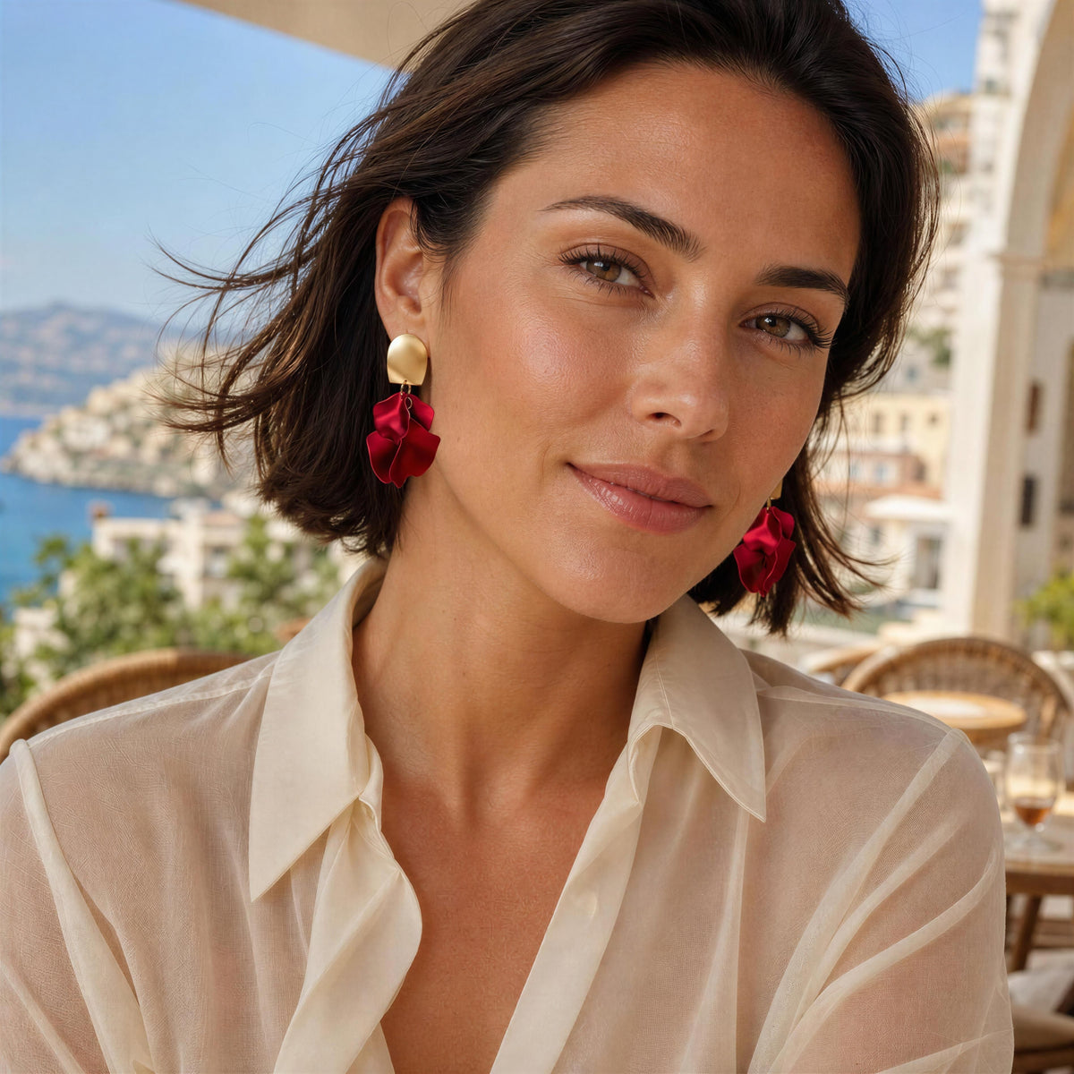 A woman wearing the Fiore Roja Earrings, a dramatic red fabric dangle earrings with gold-toned circular tops, photographed outdoors on a sunny terrace with a sea and buildings in the background.