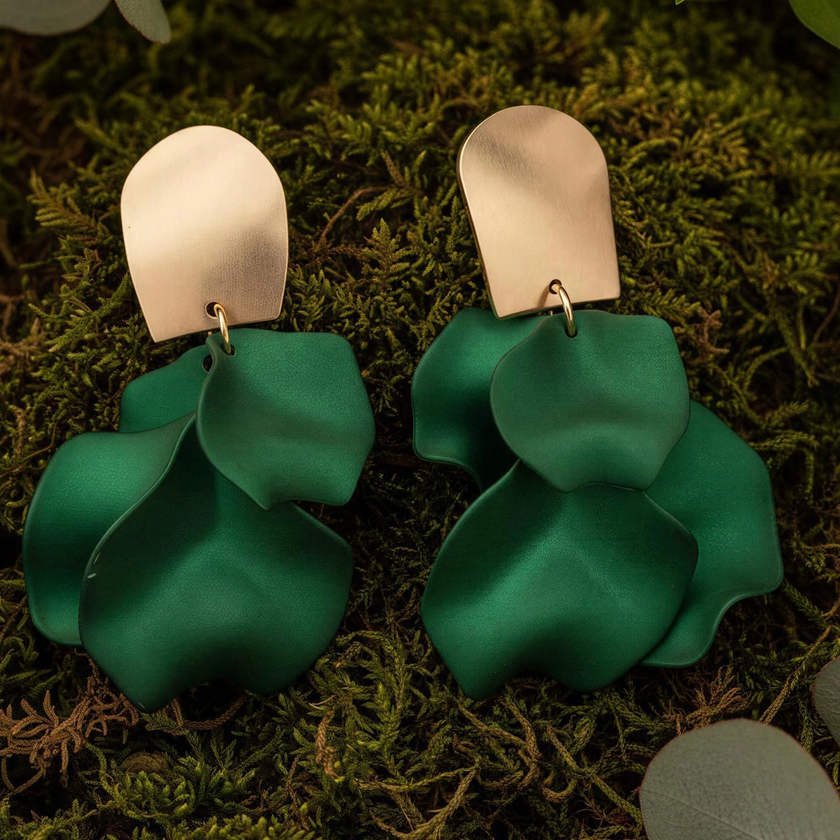 A pair of statement earrings with matte green layered petal-like drops and brushed silver-tone arch tops, displayed on a bed of green moss.