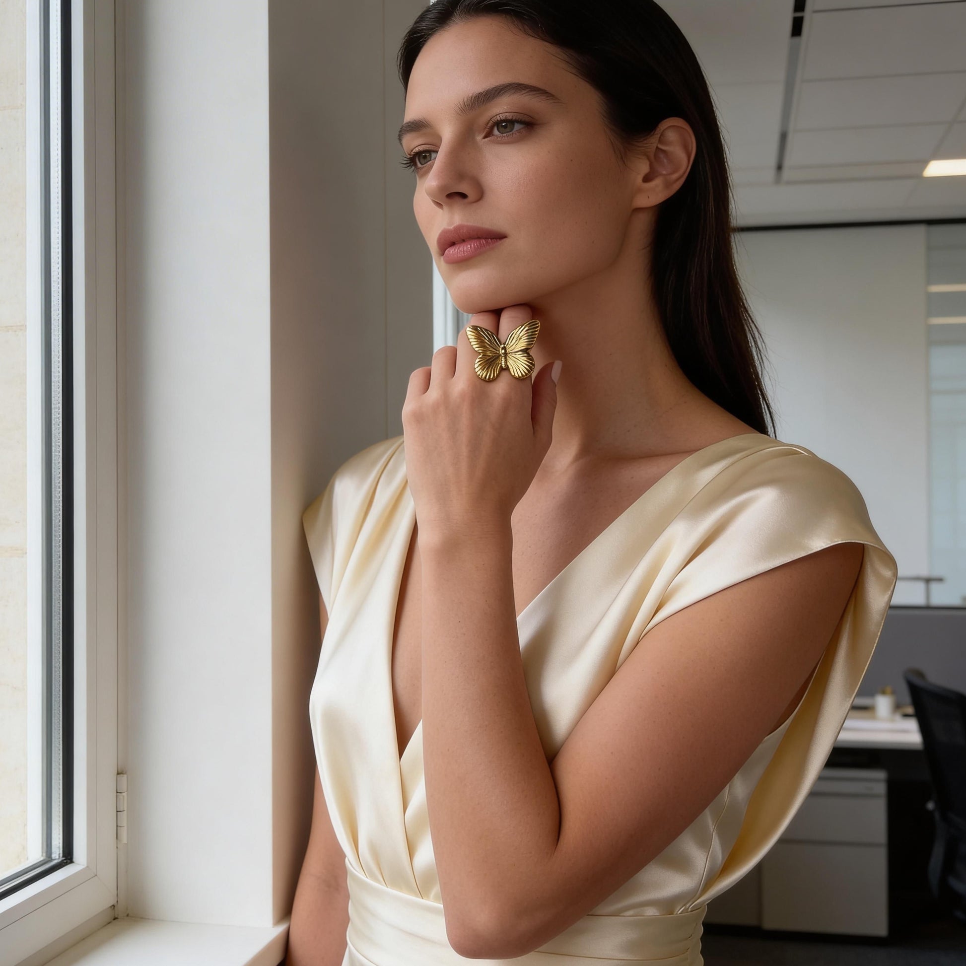Woman in a cream dress standing by a window in an office setting wearing the Forever Flutter Ring - Gold 