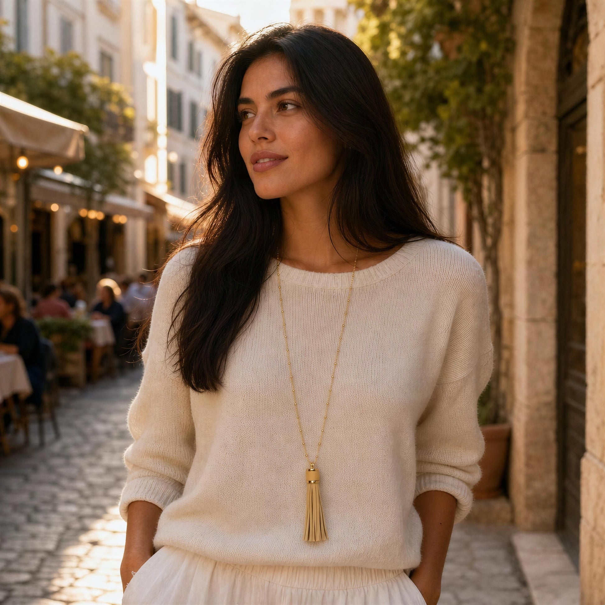 A woman with long dark hair stands on a sunlit cobblestone street wearing a cream knit sweater and a long ivory and gold tassel necklace.