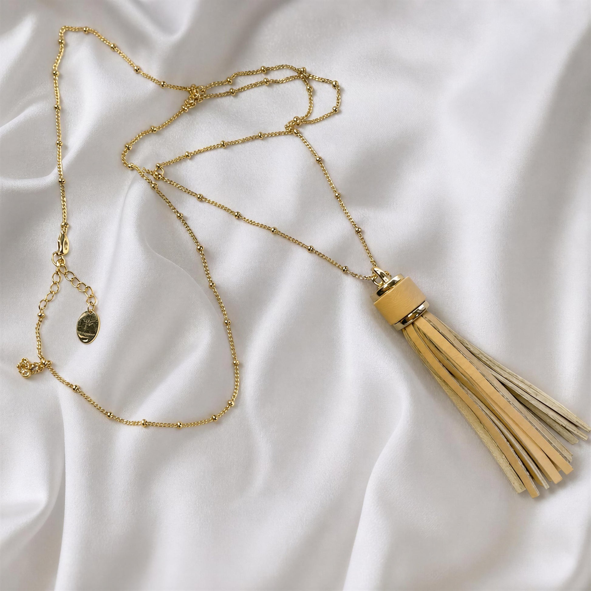 The Frangia Avorio Necklace from Chica Chica Jewelry laid out on a draped white fabric background. The pendant features a light beige wooden collar and a tassel made of multiple beige leather strands.