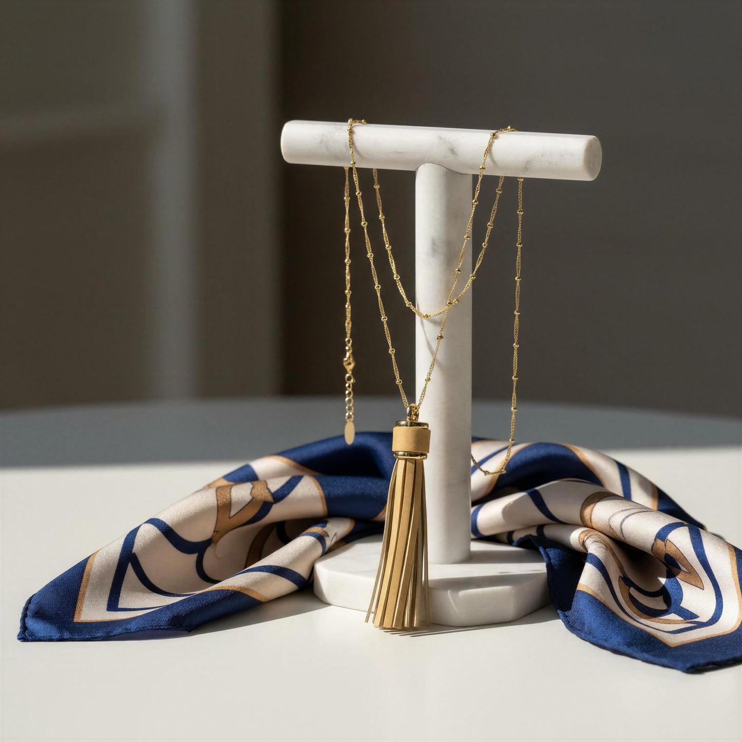 The Frangia Avorio Necklace displayed on a white marble jewelry stand, with a navy, beige, and gold patterned silk scarf draped behind it on a light surface.