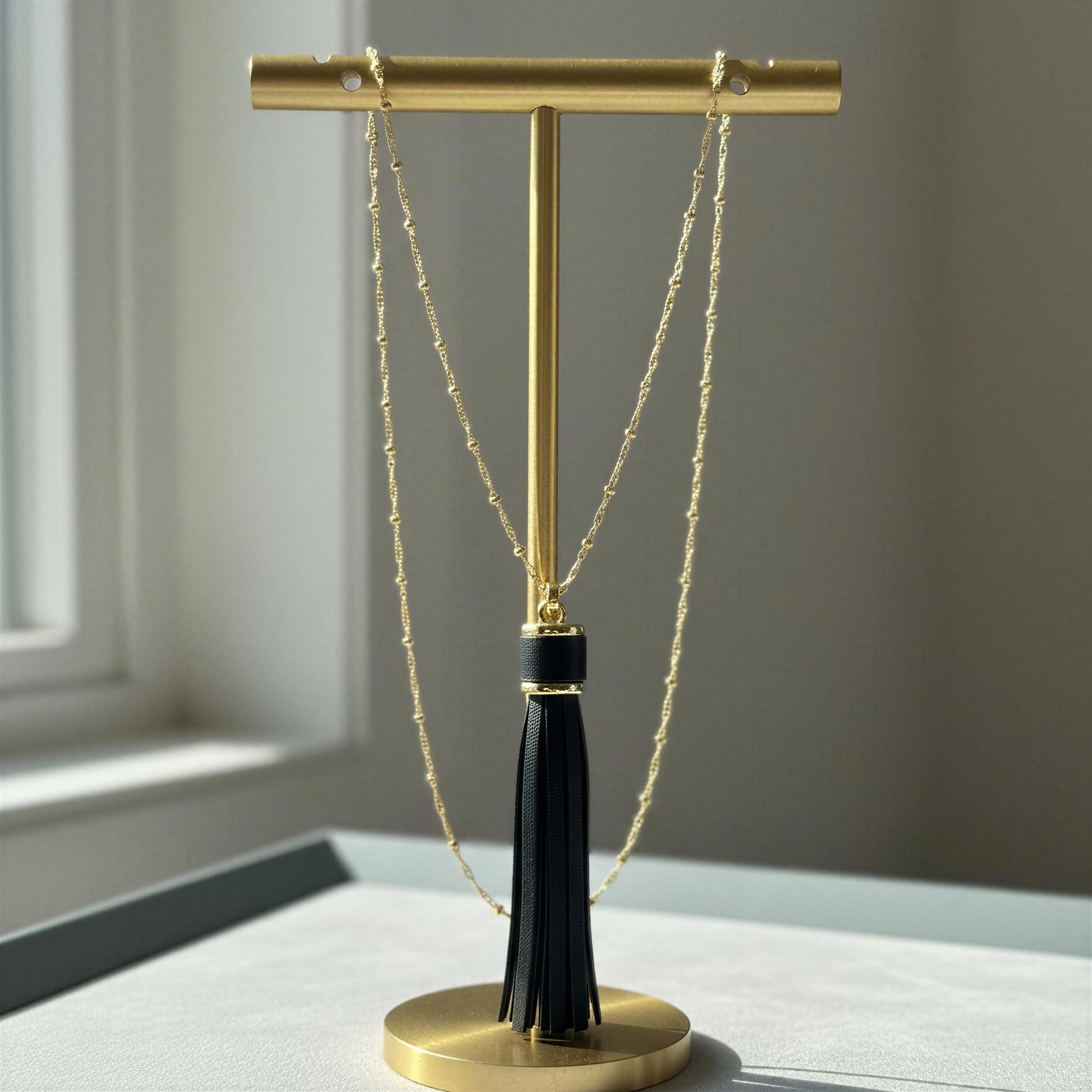 A gold T-stand jewelry display holder with a black tassel, holding the Frangia Ebano Necklace, set on a light gray surface with a soft-focus window background.