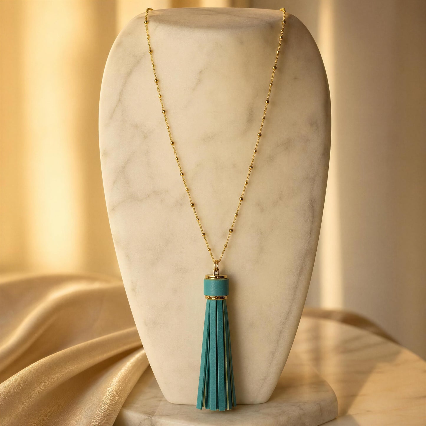 A gold-tone beaded necklace with a turquoise tassel pendant, displayed on a white marble necklace bust with draped beige fabric in the background.
