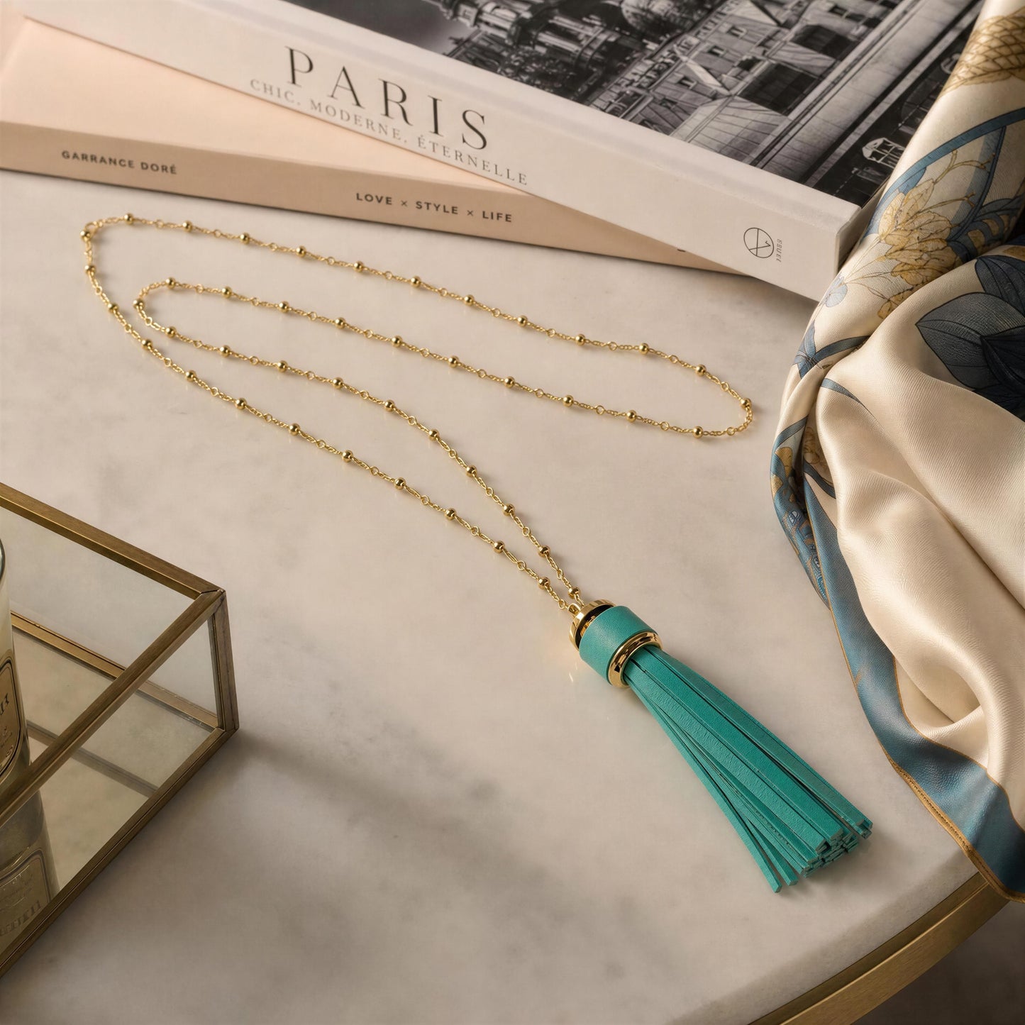 The Frangia Firuze Necklace, a gold beaded satellite chain necklace with a large turquoise tassel pendant, styled on a white marble surface with a book, a silk scarf, and a glass tray.