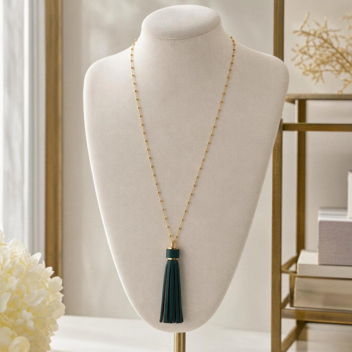 A long gold-tone beaded necklace with a dark green tassel pendant displayed on a cream jewelry bust in a softly lit indoor setting.
