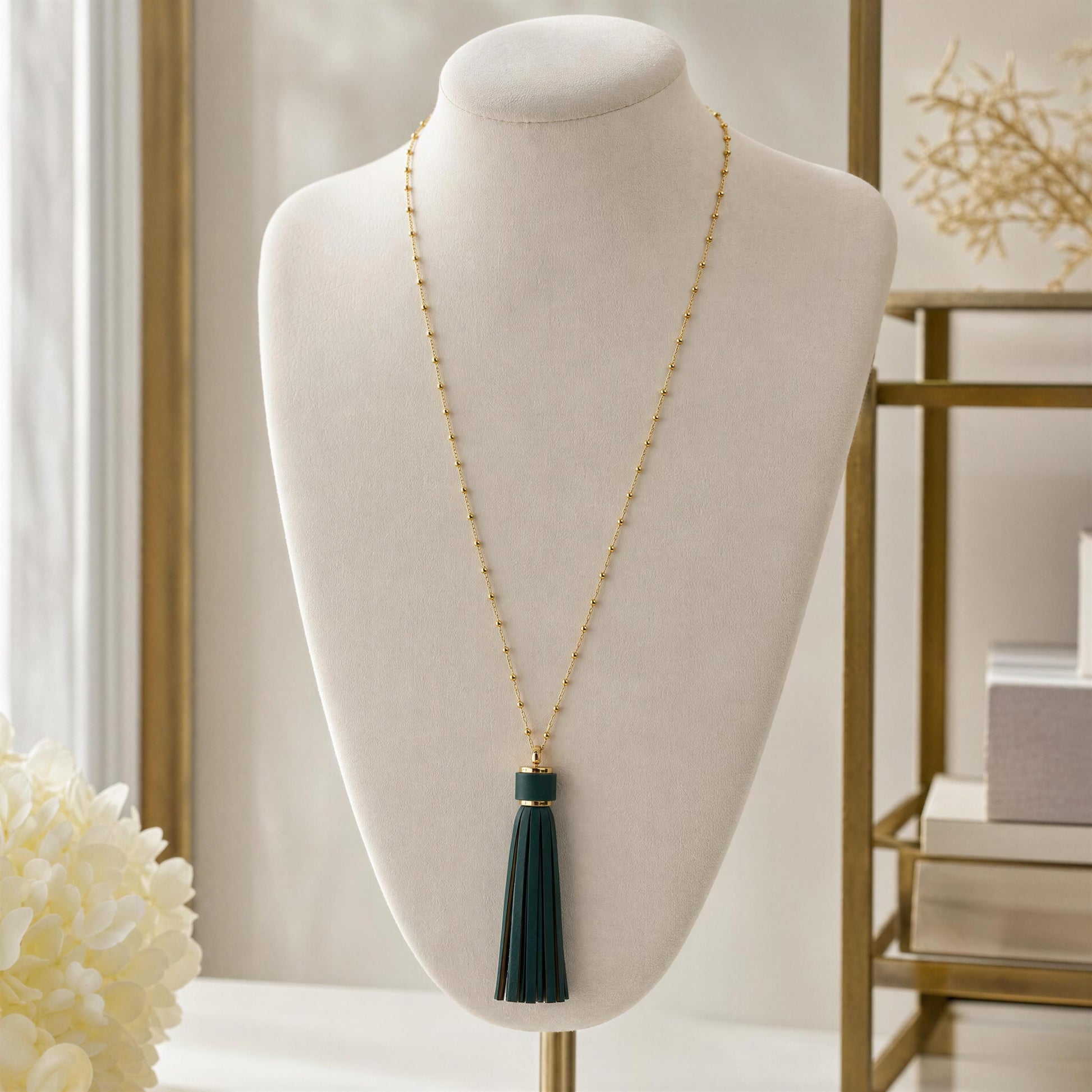 A long gold-tone beaded necklace with a dark green tassel pendant displayed on a cream jewelry bust in a softly lit indoor setting.