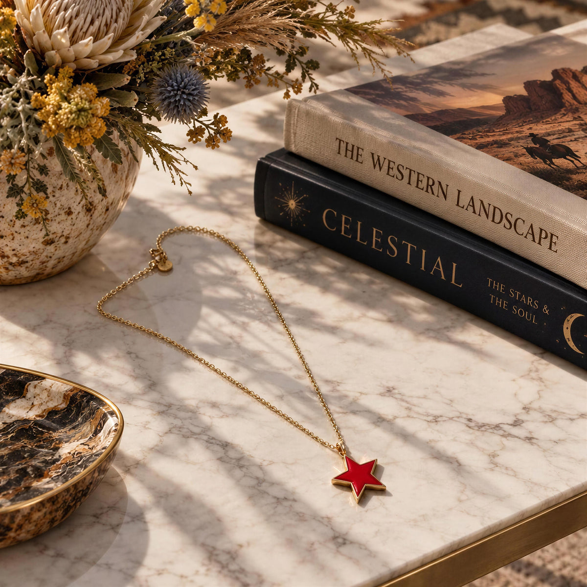 A gold chain necklace with a fuschia star pendant is styled on a white marble tabletop with dried flowers, decorative books, and a small bowl.