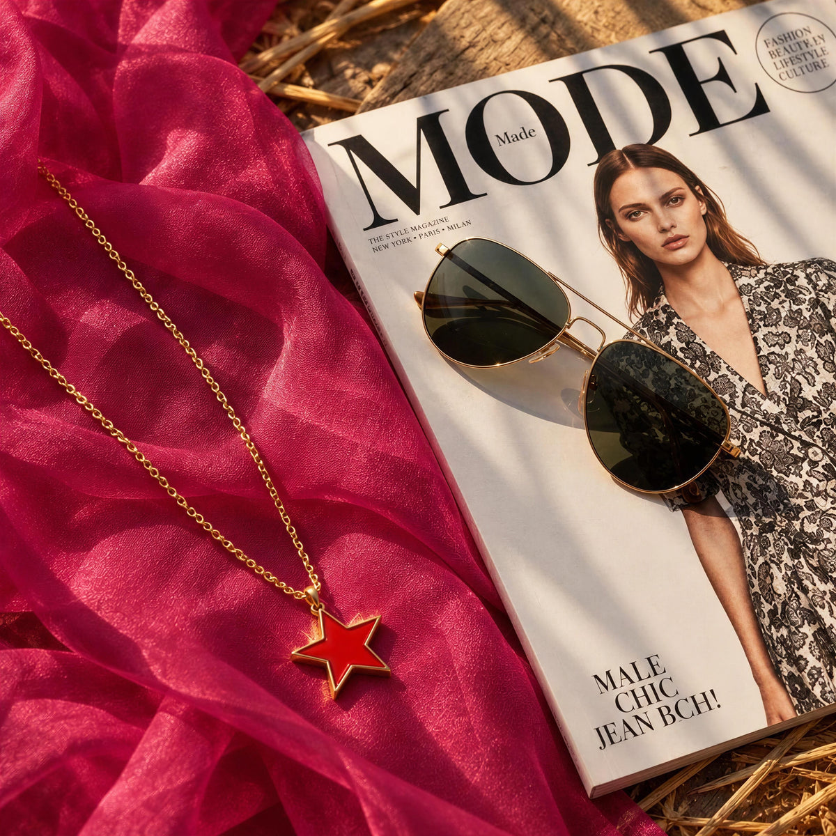 A gold chain necklace with a red star pendant is styled on bright pink fabric next to a Mode magazine and gold-framed sunglasses.