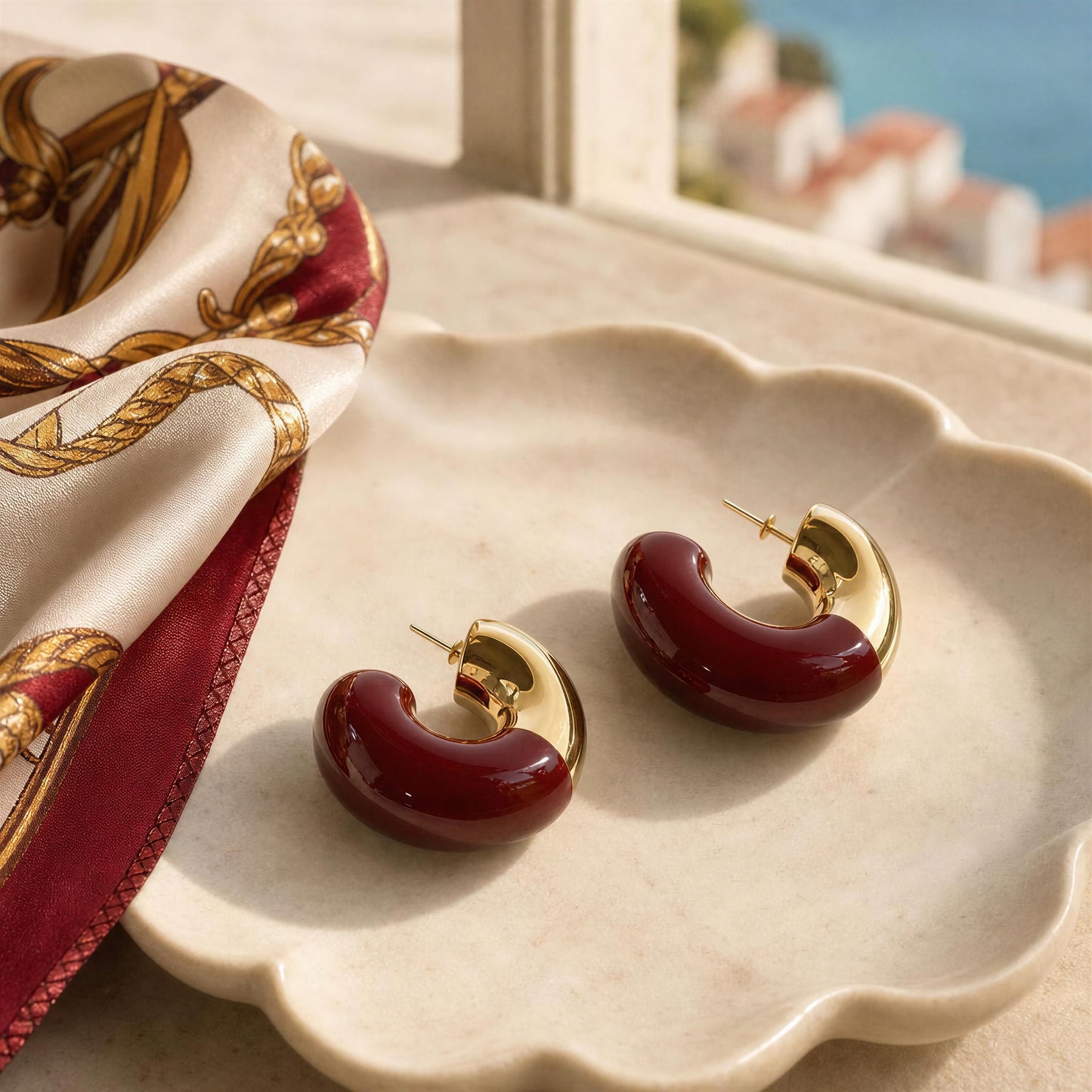 The Gatsby Arc Earrings in Merlot from Chica Chica Jewelry, displayed on a scalloped cream dish beside a patterned silk scarf, with a sunlit window and blurred coastal view in the background.