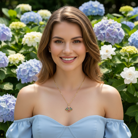 Woman wearing a blue off-shoulder top and gold Chica Chica Jewelry necklace with a green pendant, standing in front of hydrangea flowers.