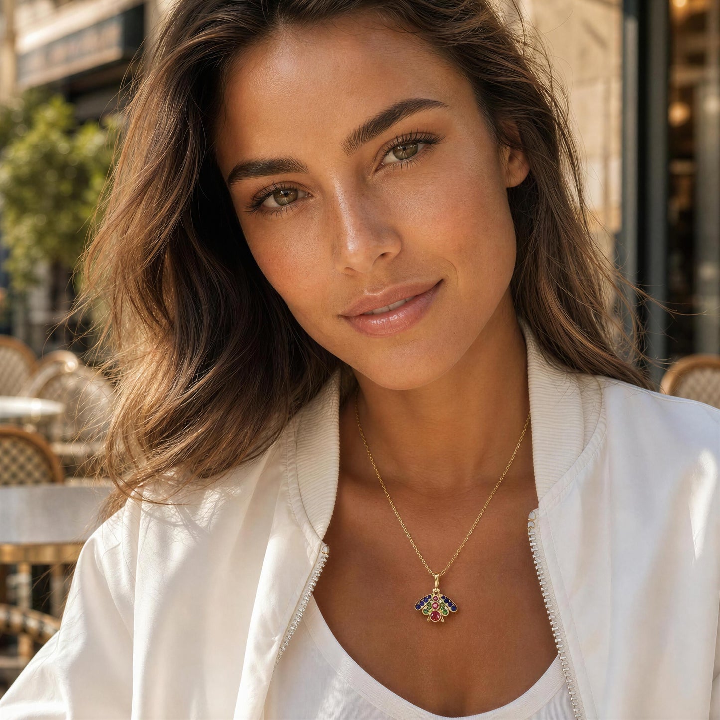 A woman wearing a delicate gold necklace with a colorful, shield-like pendant, styled with a white bomber jacket in an outdoor cafe setting.