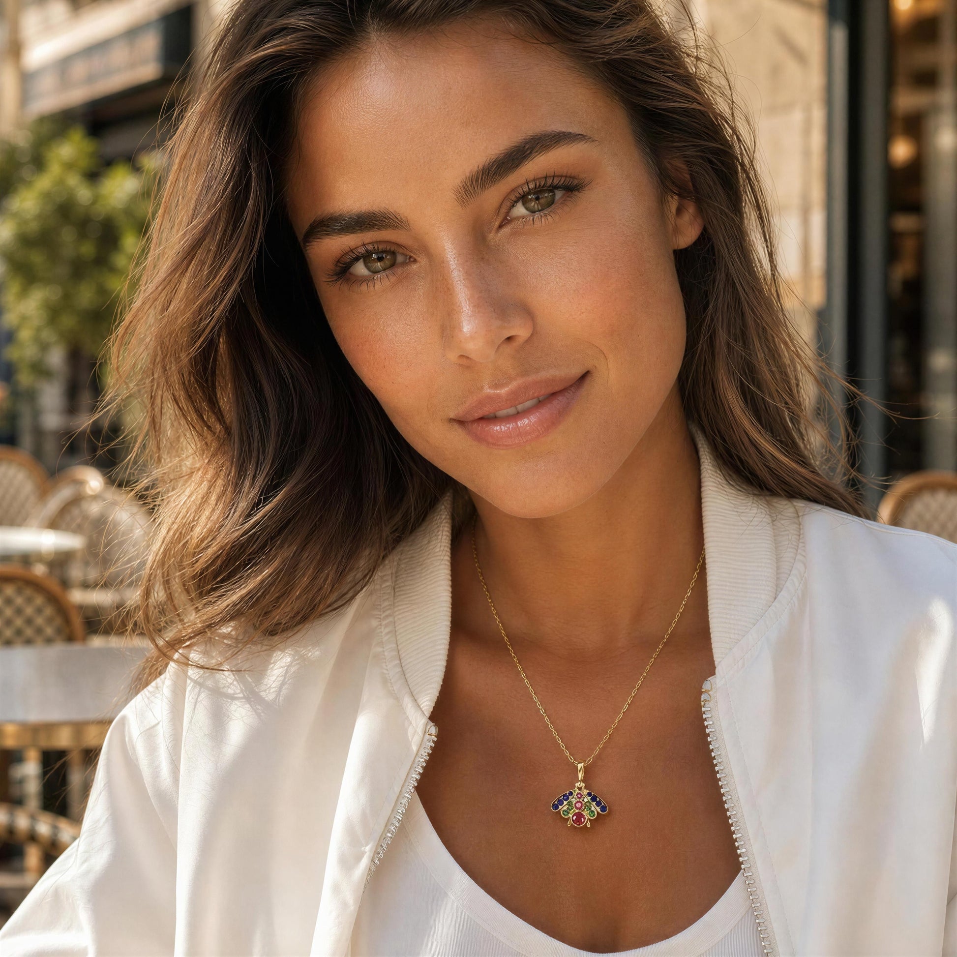 A woman wearing a delicate gold necklace with a colorful, shield-like pendant, styled with a white bomber jacket in an outdoor cafe setting.