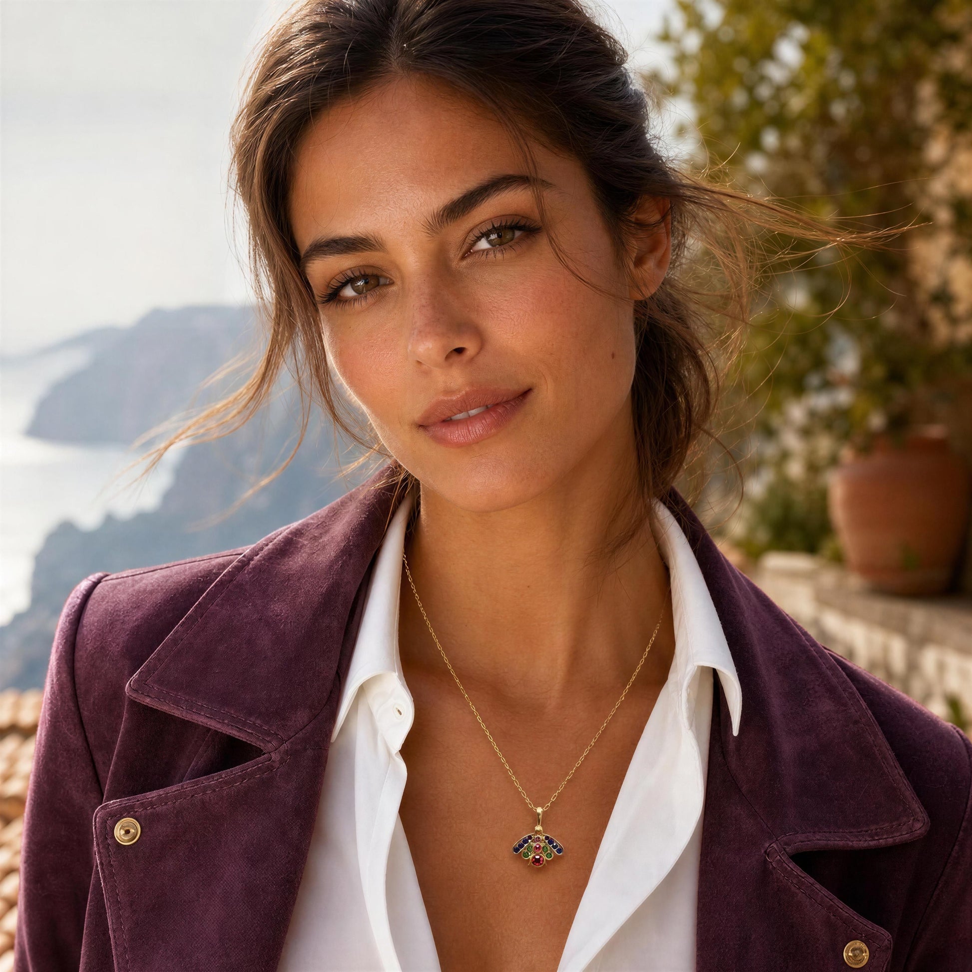 A woman wearing the Golden Meadow Bee Necklace , styled with a white collared shirt and a dark purple jacket, set against a soft-focus coastal background.